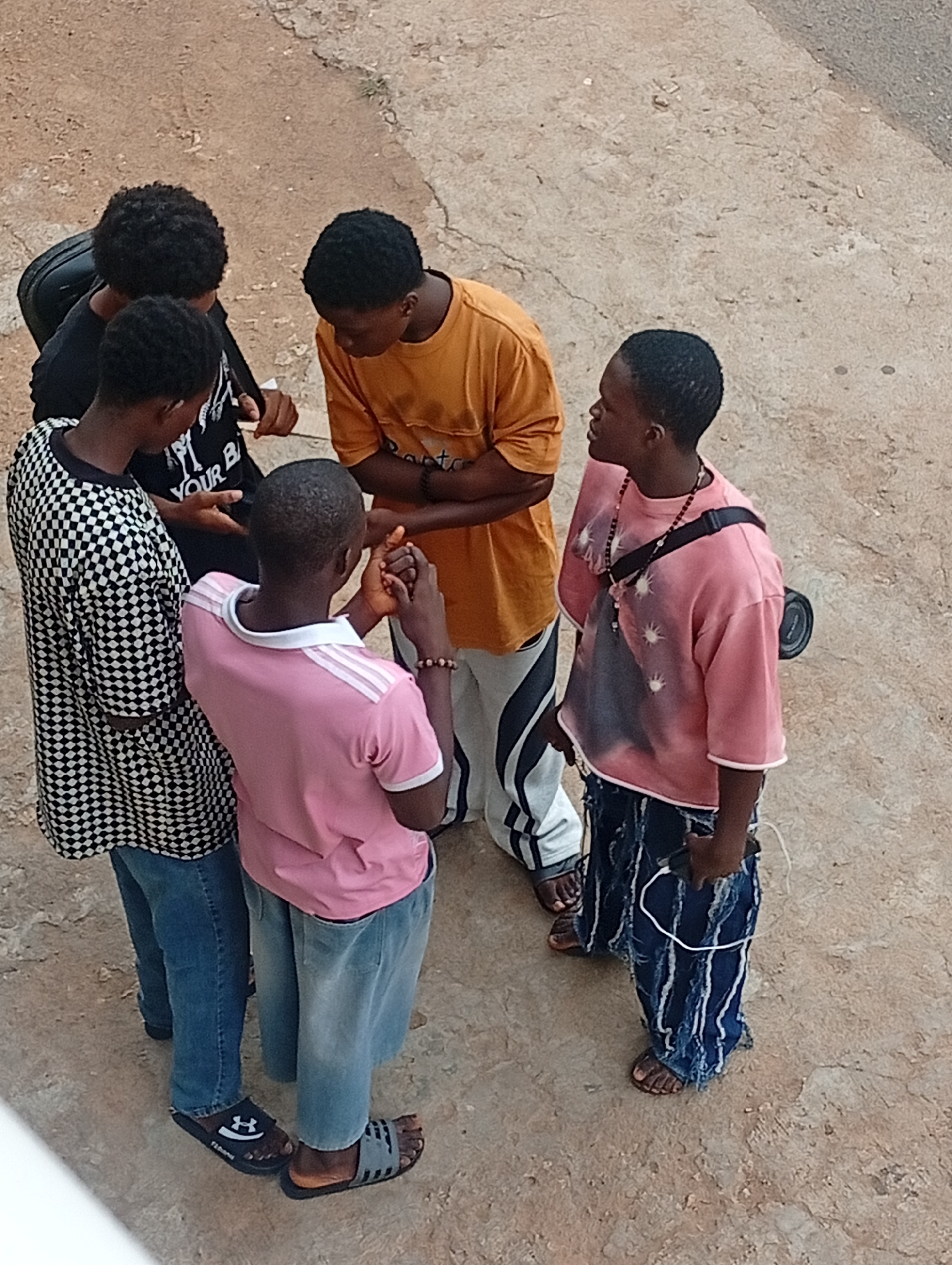 In Barakin Akawo, Nigeria, five young men are gathered in a circle on a cracked concrete surface that appears to be a public area. The individual closest to the camera is wearing a pink t-shirt with white stripes on the sleeves and light denim pants paired with dark sandals. To their left stands a person in a black and white checkered shirt and jeans, also wearing sandals. One man, facing away from the camera, wears a black t-shirt. A man in an orange t-shirt is visible to the right and stands with his arms crossed. Another man wears a pink t-shirt, blue jeans and has a camera strapped to his side. The group appears to be engaged in conversation, with some holding or gesturing with their hands. One of the men is holding a white phone. The setting suggests an outdoor urban environment.