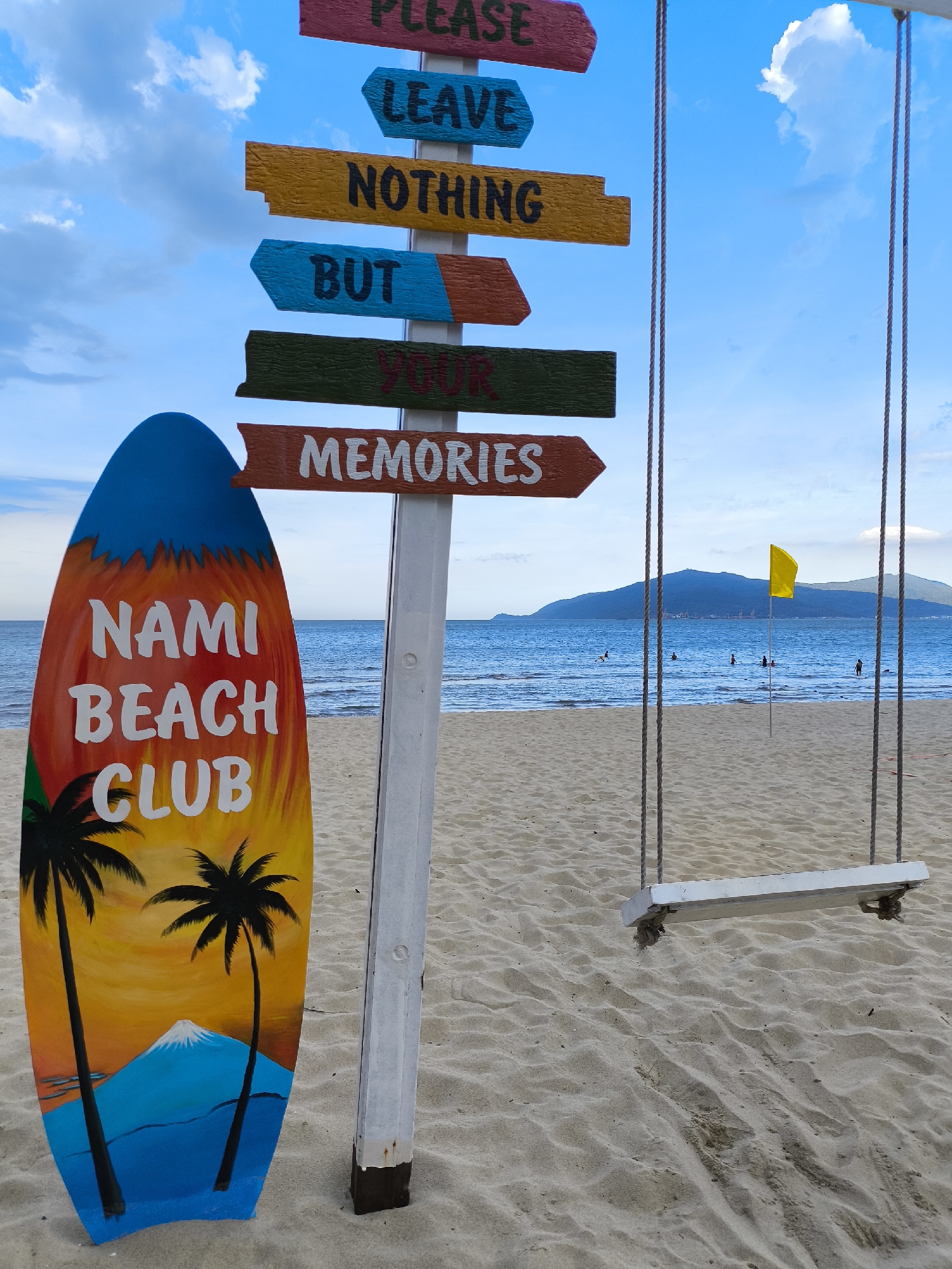 On a sandy beach, a surfboard with the words "Nami Beach Club" painted on it stands upright. To the right of the surfboard is a wooden signpost with multiple colored arrows pointing in different directions. The top arrow is red and reads "PLEASE". The second arrow is blue and reads "LEAVE". The third arrow is yellow and reads "NOTHING". The fourth arrow is blue and reads "BUT". The fifth arrow is green and appears to have faded writing, possibly "YOUR". The bottom arrow is orange and reads "MEMORIES". In the background, the ocean stretches out to the horizon with a distant island. Several people are visible in the water. A yellow flag is planted in the water near the shore. A swing set with a white wooden seat hangs from ropes on the right side of the frame, partially obscuring the view of the ocean and island. The scene suggests a tropical beach destination.