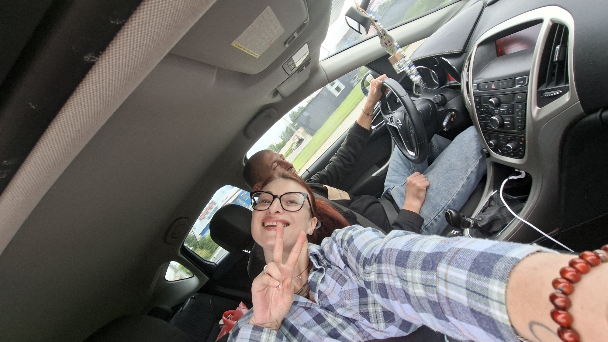 An outdoor, wide-angle, eye-level shot shows two individuals inside a car. The driver, a man with short hair and a beard, is wearing a black hoodie and jeans. He is looking forward and has his hands on the steering wheel. Beside him, in the passenger seat, a woman with red hair and glasses is looking at the camera and making a peace sign with her hand. She is wearing a blue and white plaid shirt and jeans. The car's interior is visible, including the dashboard, steering wheel, and gear shift. The background shows a street with buildings and trees. The weather appears to be overcast. The location is identified as Smyalavichy, Belarus.