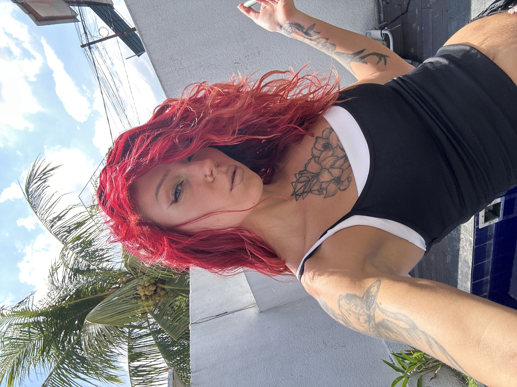 A woman with bright red, wavy hair and tattoos is taking a selfie outdoors. She is wearing a black tank top with a white underlayer. She has a nose ring and is holding a cigarette in her right hand, though it does not appear to be lit. Behind her, a palm tree with coconuts and a white wall are visible. The sky is blue with scattered clouds. The location is identified as North Kuta, Indonesia.
