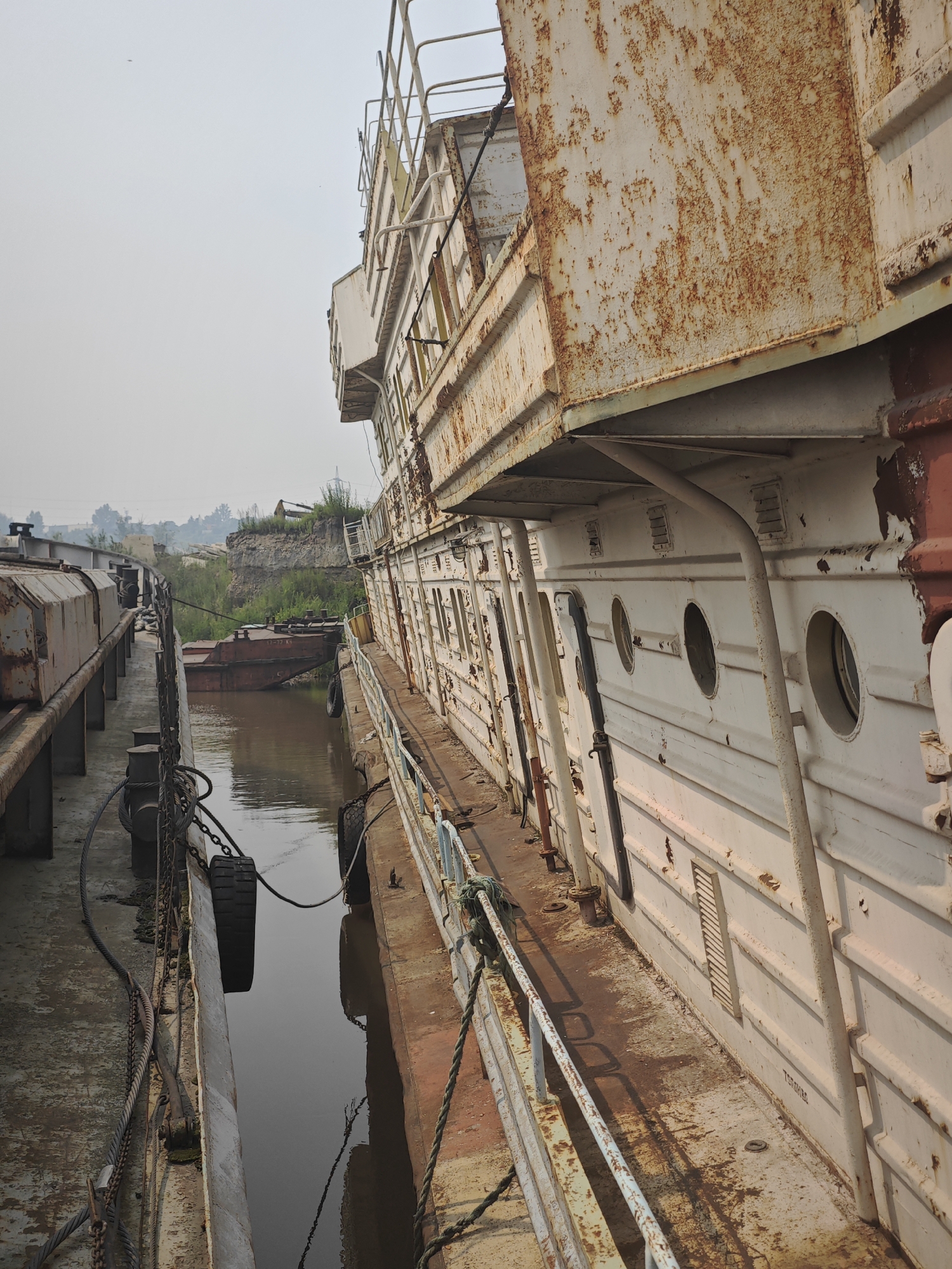 The image depicts a close-up, eye-level view of two weathered vessels moored side-by-side in a narrow waterway. The vessel on the right, largely filling the frame, is white with extensive rust-colored streaks and patches, particularly on its upper structures and along the hull. It features rows of circular portholes and rectangular vents. A metal railing with a weathered appearance runs along its deck. The vessel on the left is partially visible, appearing more utilitarian and metallic. The waterway between the vessels is calm and reflects the overcast sky. In the background, a steep, vegetated embankment rises from the water, with a disused piece of heavy machinery visible at the top. The overall impression is one of decay and abandonment, possibly in an industrial or port area. Based on an understanding of common imagery associated with Krasnoyarsk, Russia, this scene is likely located on a branch of the Yenisei River or one of its tributaries near the city, where older vessels are often moored or decommissioned. No people are visible in the image.