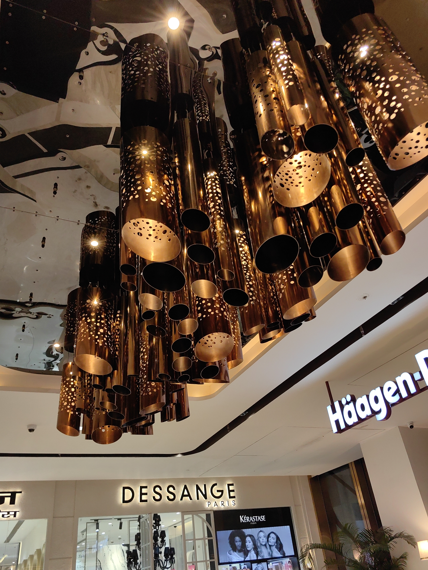 The image depicts an interior view of a modern shopping mall in Mumbai, India. The foreground is dominated by a large, elaborate chandelier made of numerous interconnected cylindrical metallic tubes, some of which are perforated with circular patterns, casting warm light. The ceiling is highly reflective, likely a mirrored surface, showing distorted reflections of the chandelier and the mall's architecture.
Below the chandelier, visible signs indicate retail establishments. On the left, a sign in a non-English script (likely Hindi or a related Indian language) is partially visible, followed by a sign that reads "DESSANGE PARIS". To the right of "DESSANGE PARIS", a sign for "Häagen-Dazs" is clearly visible. Beneath the "DESSANGE PARIS" sign, a display screen shows an advertisement featuring four women, and the text "KERASTASE PARIS" is also present.
The overall environment appears to be well-lit and contemporary, characteristic of a high-end retail space. No people are distinctly visible in the immediate foreground or midground, though the reflections in the ceiling suggest the presence of other shoppers or elements within the mall. The perspective is upward-looking, emphasizing the grand scale of the chandelier and the mall's ceiling design.