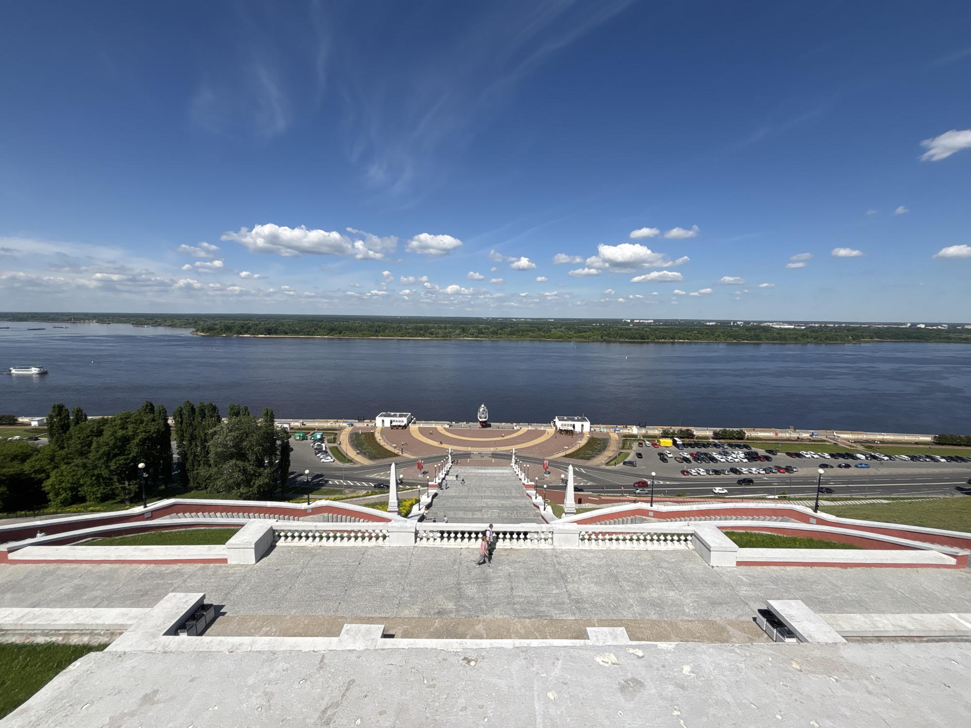 A high-angle, wide-angle photograph captures a panoramic view overlooking a grand, multi-tiered staircase descending towards a wide river under a bright blue sky. The image was taken on a sunny day with scattered white clouds, suggesting pleasant weather.
In the foreground, the upper sections of a monumental stone staircase are visible, featuring wide steps, red retaining walls, and white balustrades. Several people are seen walking down the stairs, appearing as small figures due to the distance. The staircase narrows as it descends, leading to a semi-circular promenade at the river's edge.
At the base of the staircase, the promenade is paved and features a central monument or fountain. Two small, white buildings with dark roofs flank this central feature. A road runs parallel to the river behind the promenade, with a large parking lot filled with numerous vehicles visible to the right. Green grassy areas are interspersed along the sides of the staircase and the riverbank. Tall, dark green trees line parts of the embankment on the left.
The wide river, likely the Volga or Oka given the Nizhny Novgorod context, occupies the middle ground of the image. Its deep blue surface reflects the sky. A white passenger boat or ferry is visible on the water to the far left. The opposite bank of the river is heavily forested, with a distant low-rise urban skyline barely discernible on the horizon. The overall scene depicts a scenic public space, likely a popular vantage point or recreational area. No discernible text is visible in the image.