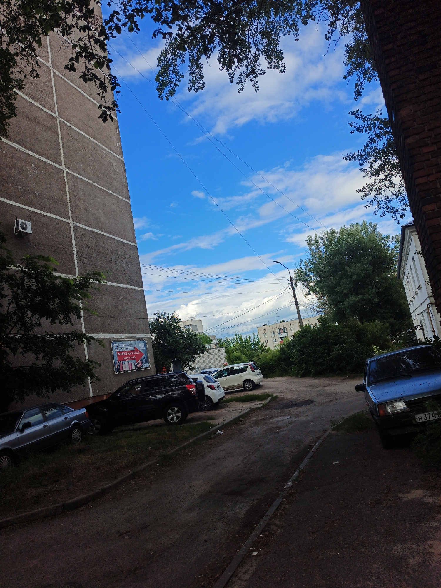 The image depicts a street scene in Kursk, Russia, likely in a residential area. A multi-story apartment building with a textured, grey facade dominates the left side of the frame, with vertical white stripes marking its structure. Overhanging tree branches with green leaves frame the top and left portions of the image. A blue sky with scattered white clouds serves as the background.

Several cars are parked on a dirt and asphalt lot adjacent to the building. These include a silver sedan on the far left, a black SUV in the middle, and a white hatchback behind it. A blue sedan is parked on the right side of the lot, its license plate partially visible.

Further in the background, a street lamp is visible, along with more trees and buildings, suggesting an urban environment. The overall impression is of a typical day in a Russian city courtyard.