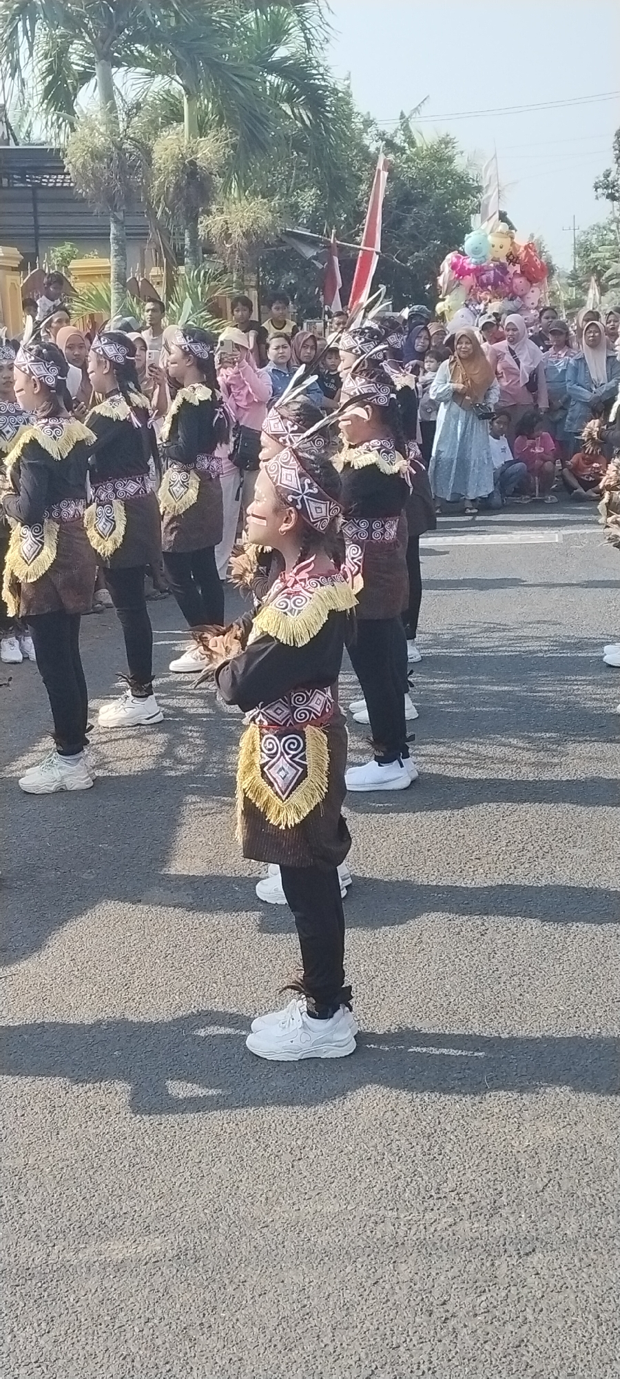 This is a medium shot, eye-level, outdoor photograph taken in the city of Yosowilangun Kidul, Indonesia. The image features a group of young people in traditional attire marching on a paved road. The individuals are wearing black long-sleeved tops adorned with intricate cultural patterns in white, red, and gold, and brown skirts with decorative fringe. They also have matching headbands and feathers in their hair, and black pants paired with white athletic shoes. Some are holding objects in their hands, possibly for a performance.
In the background, there is a crowd of spectators watching the procession. Some are holding phones, suggesting they are capturing the event. There are also palm trees and buildings visible, indicating an urban setting. The scene appears to be a cultural parade or festival. The lighting suggests it is daytime, with clear skies.