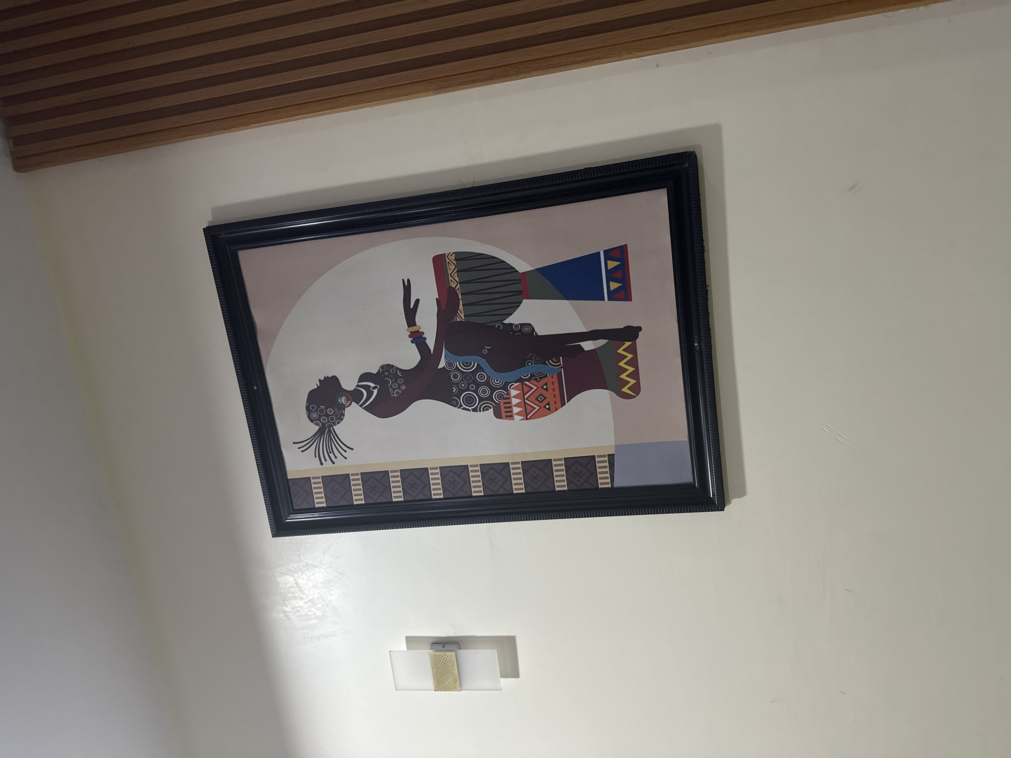 The image displays an interior scene featuring a framed artwork prominently mounted on a light cream-colored wall. The artwork, enclosed in a black frame with decorative molding, depicts a stylized figure of an African woman in a reclining or supine posture. She is adorned with intricate circular and geometric patterns on her body or clothing, and wears bangles on her raised right arm. Alongside her, a tall, djembe-like drum is shown with vibrant geometric patterns in blue, red, and yellow on its base. The background of the painting features a large light-colored arch and a patterned horizontal strip at the bottom.
Above the wall, a slatted wooden ceiling with light brown planks is visible. Below the artwork, a small, modern wall-mounted light fixture is affixed to the wall. This fixture consists of a metallic central component flanked by two rectangular, translucent light diffusers. The lighting in the room appears to be artificial, casting some reflections on the wall's surface. The image is taken at an angle, causing the entire composition to appear tilted. No specific activity is taking place other than the display of the artwork and the ambient room setting. There is no visible text in the image.