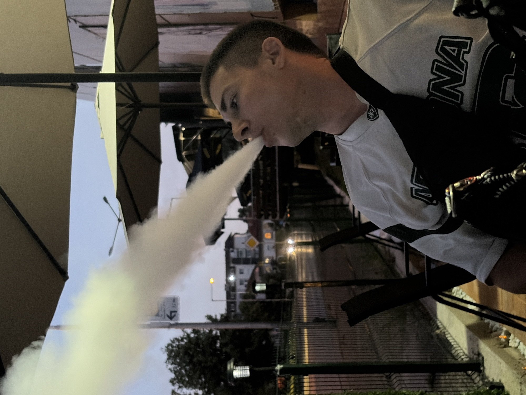 A young Caucasian male is exhaling a large plume of white smoke, likely from a hookah or vape, in an outdoor setting at dusk. He is seated at a table, possibly at a cafe or restaurant, with an umbrella providing shade overhead. He is wearing a white long-sleeved shirt with black accents and lettering across the chest, and a black crossbody bag. In the background, a street is visible with cars and buildings, suggesting an urban environment. The lighting indicates it is either early morning or late evening.