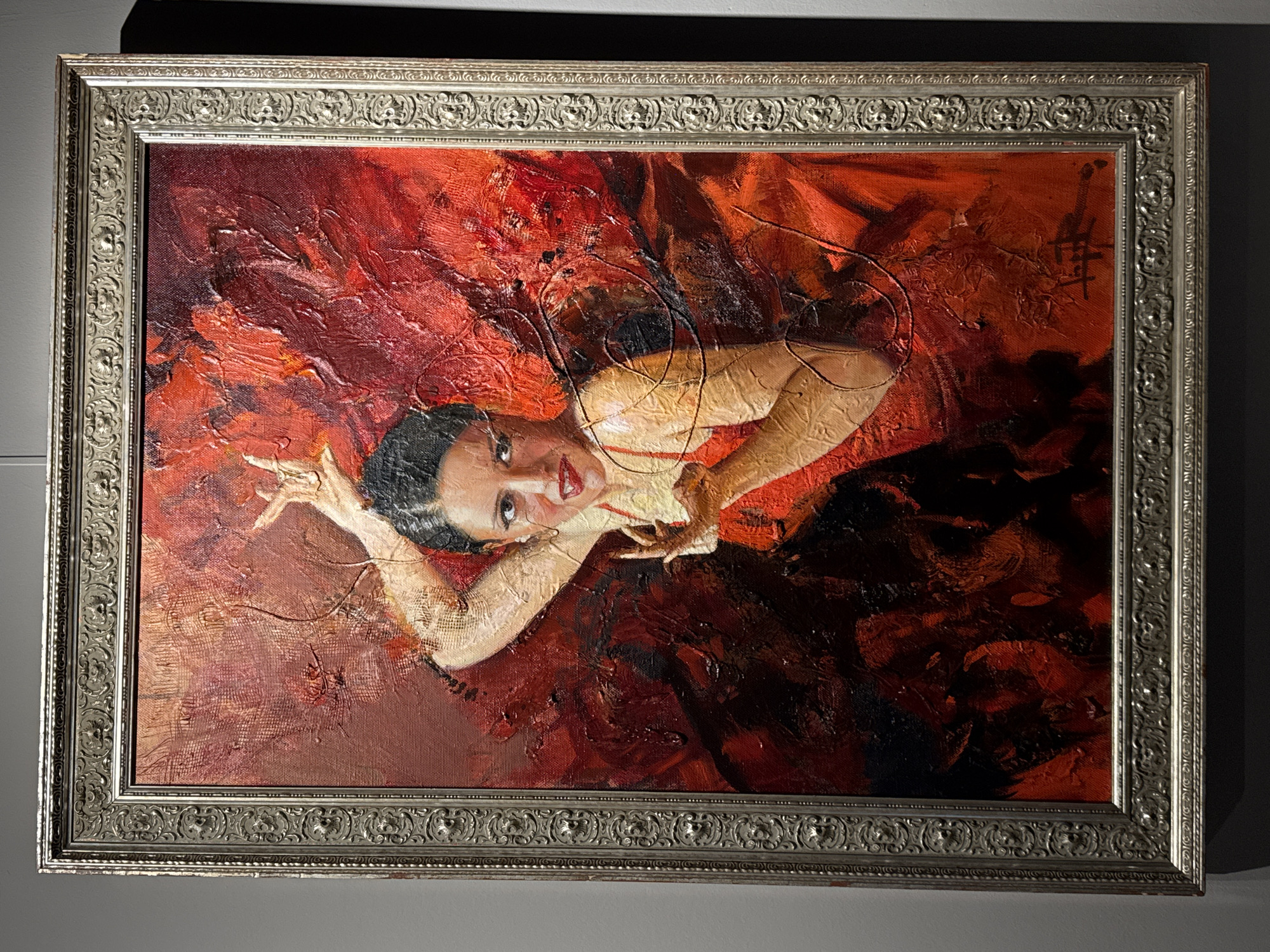 A framed oil painting on canvas depicts a woman dancing flamenco. The painting is in the style of a figurative expressionist work, with bold brushstrokes and a vibrant palette dominated by reds and blacks. The woman, with dark hair pulled back, is wearing a red dress and her arms are raised and her hands are in mid-gesture. Her face is turned towards the viewer with a focused expression. The background is abstract and energetic, with swirling textures and colors that convey movement and passion. The painting is housed in a decorative silver-toned frame with intricate detailing. The artwork is displayed against a plain gray wall, suggesting it is in a gallery or exhibition space. The image does not provide any specific information about its location in Saint Petersburg, Russia.
