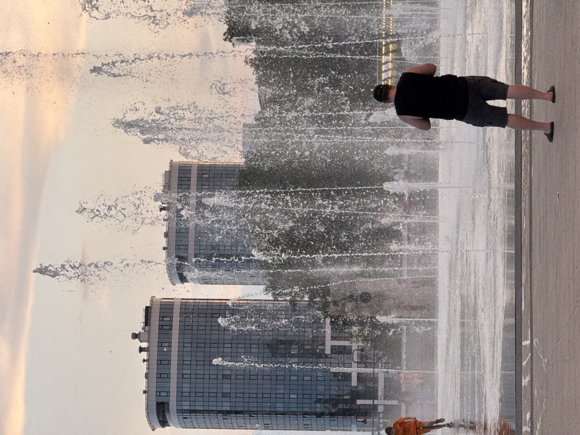The image depicts a wide public fountain in the city of Izhevsk, Russia. Two modern glass-facade buildings are visible in the background, suggesting an urban setting. In the foreground, a young boy wearing an orange shirt and shorts is running across the wet ground near the fountain, likely playing. Further back, a man in a black sleeveless shirt and gray shorts stands facing the fountain, with his back to the camera. The fountain itself is active, with numerous jets of water creating a misty spray. The sky is a pale orange and pink, indicating either sunrise or sunset. The ground around the fountain appears to be paved, and there are trees and other people blurred in the background.