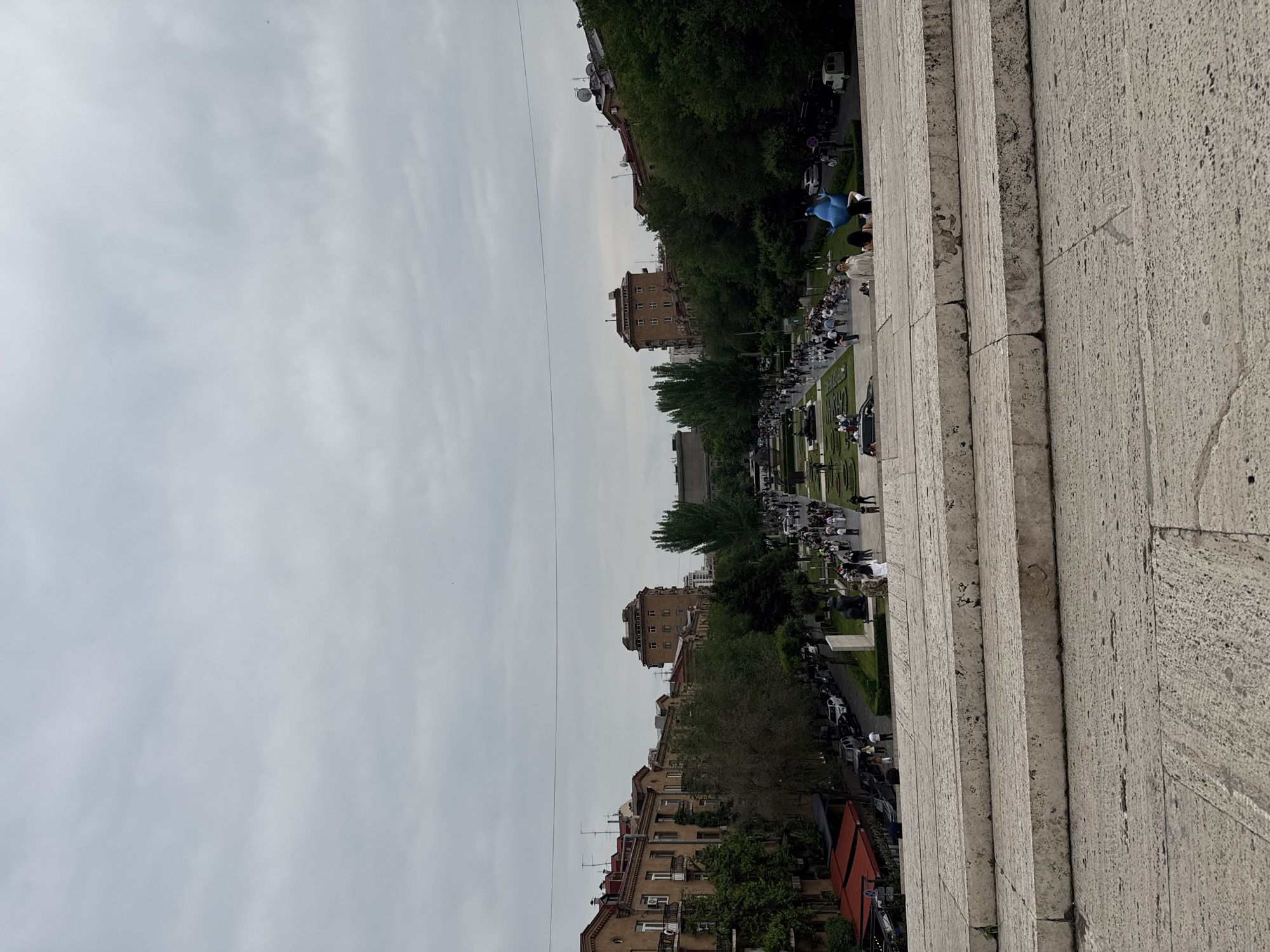 The image depicts a view of Yerevan, Armenia, likely from a high vantage point on a stone structure, possibly a staircase. The sky is overcast. Below, a park stretches into the distance, bordered by trees and buildings of various heights and architectural styles. People are walking along a path within the park. Some are gathered in small groups. Cars are parked on the sides of the street. The buildings are generally beige or brown with red-tiled roofs. Power lines run across the sky.