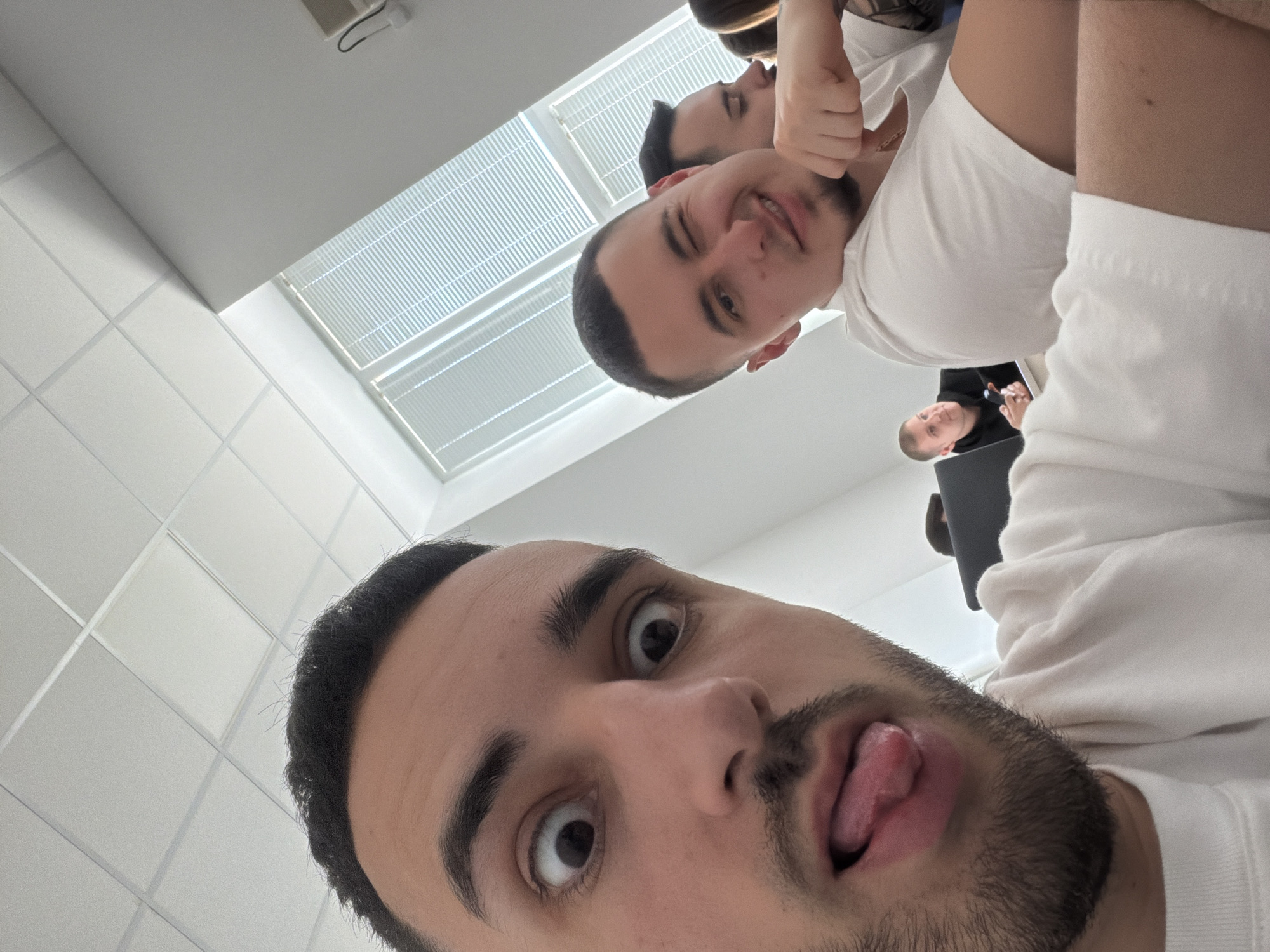A close-up selfie of a man with dark hair and a beard, sticking his tongue out. Behind him, slightly out of focus, are two other men. One man is smiling and holding his fist up to his face, while the other man behind him is looking towards the window. All three men are wearing white t-shirts. In the background, there is a window with blinds and a white tiled ceiling. The location is likely indoors but the location in Plovdiv, Bulgaria cannot be determined from the image.