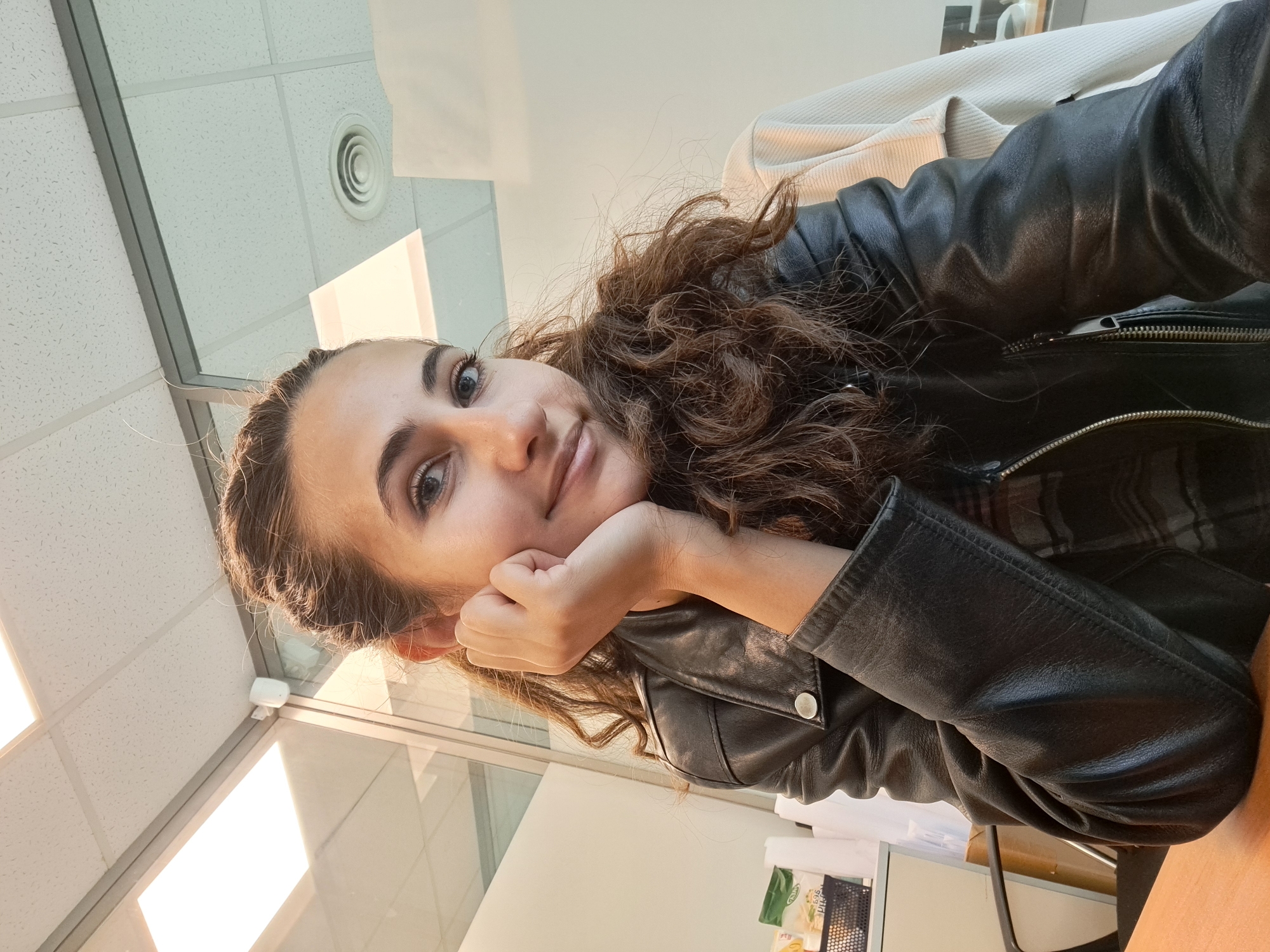 A young woman with long, curly brown hair is captured in a close-up, indoor, selfie-style photograph. She is seated, with her left hand resting on her chin and her head tilted slightly. She wears a black leather jacket over a plaid shirt. Her expression is pleasant, with a gentle smile and direct eye contact with the camera. The background suggests an office environment, with a drop ceiling with fluorescent lighting panels, glass partitions, and office furniture visible. The lighting is bright, casting soft shadows.