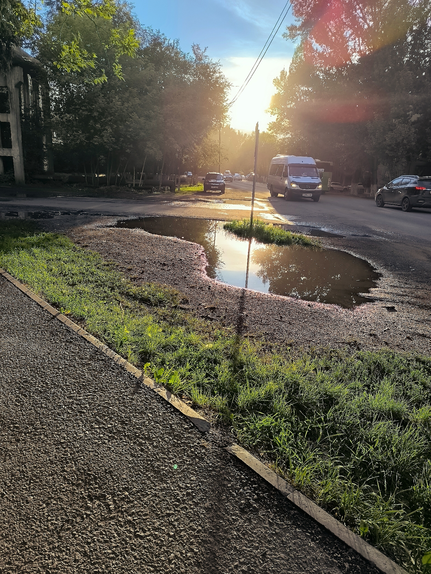 A low-angle, full shot captures a sun-drenched street scene in the city of Qarağandı, Kazakhstan. A large puddle dominates the foreground, reflecting the golden afternoon sun and the surrounding trees. To the left of the puddle is a strip of gravel, bordered by a concrete curb and lush green grass. Beyond the puddle, a paved road stretches into the distance, with a white van and several other cars driving on it. The sun's rays beam through the trees on the right side of the image, creating lens flare and a warm, hazy atmosphere. The environment appears to be a suburban or semi-urban area, possibly near a park or residential zone. The lighting suggests late afternoon or early evening.