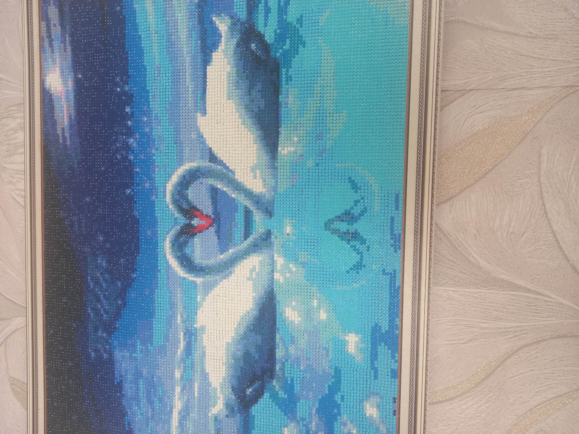 The image displays a framed diamond art painting of two white swans with their necks intertwined, forming a heart shape. The swans are depicted in a body of water, with their reflections visible below. The background is a dark blue sky with stars. The painting is framed in a silver and gold patterned frame and hangs on a wall with textured wallpaper. The location of the image is in Komsomolskoye, Russia.