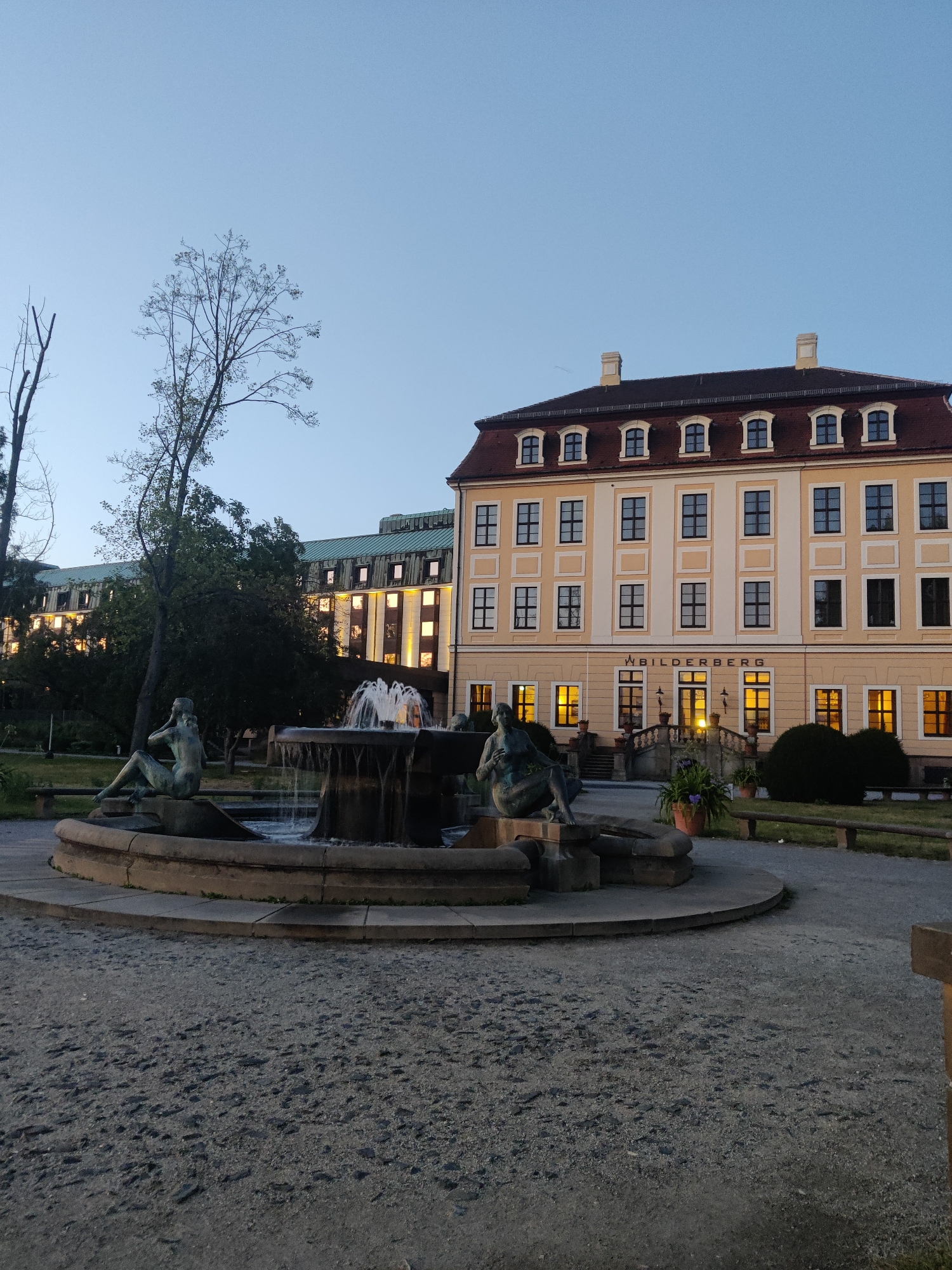 The image depicts a twilight scene at a hotel in Dresden, Germany, identified by the signage on the building as "BILDERBERG." In the foreground, a stone fountain with sculptures of reclining figures is active, with water cascading into the basin. The fountain is surrounded by a gravel pathway and manicured landscaping.
Behind the fountain, the hotel building is visible. It is a light-colored, multi-story structure with a dark red roof and dormer windows. The hotel's main entrance features a set of stairs leading up to a double door. The windows of the hotel are illuminated with warm, yellow light, suggesting interior lighting. The hotel's facade is adorned with decorative architectural elements.
To the left of the hotel, another, larger building with a green roof and multiple rows of illuminated windows is partially visible, indicating it might be an adjacent structure or an extension of the hotel complex. Several mature trees are present in the scene, some bare-branched and others with foliage, contributing to the park-like atmosphere. The sky is a clear, pale blue, indicative of the late evening or early morning hours. The overall impression is one of a serene and well-maintained public space associated with the hotel.