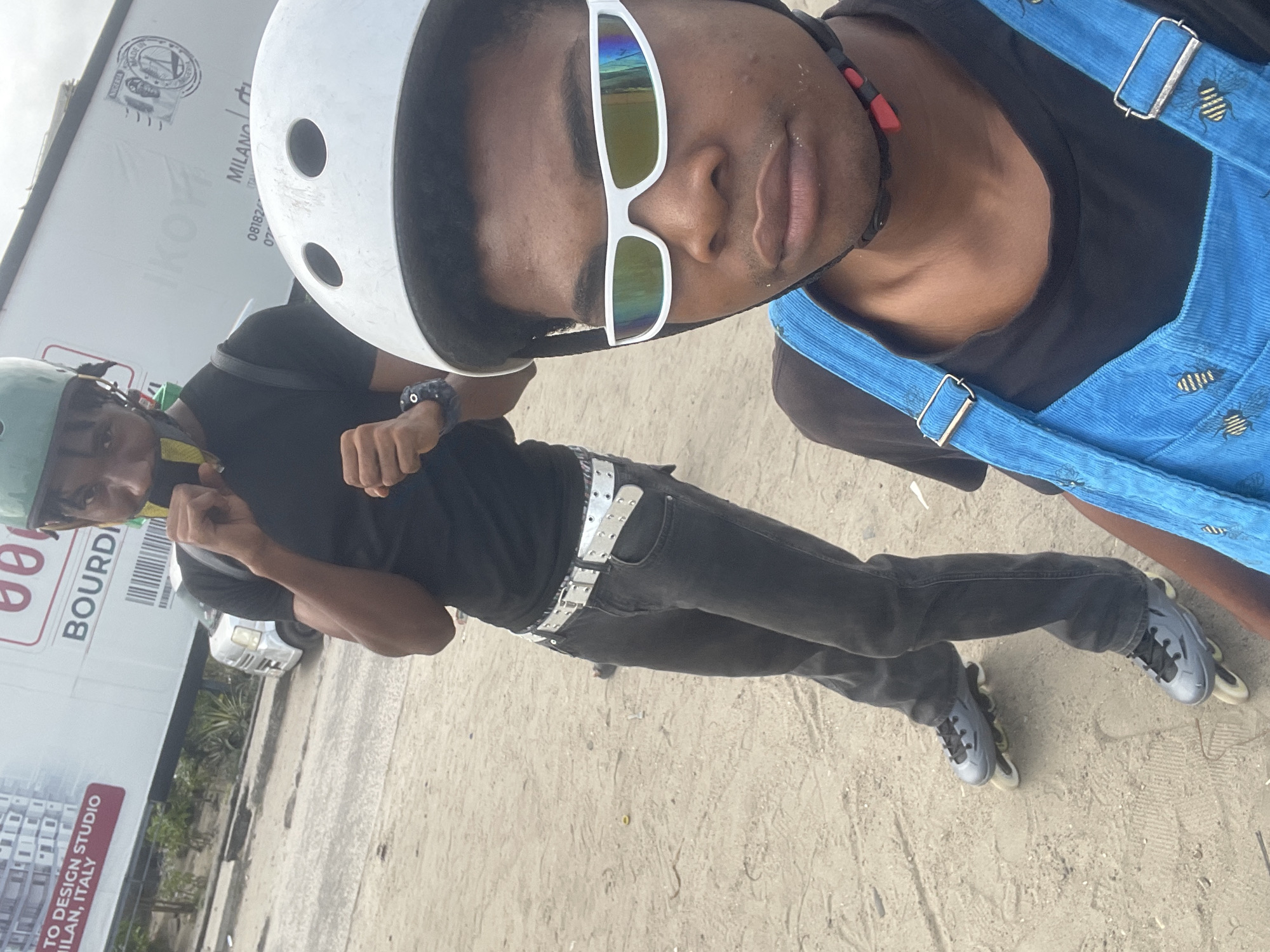 The image shows two young Black individuals in Ilado, Nigeria, engaged in rollerblading. The person in the foreground is wearing white futuristic-style sunglasses, a white helmet, and a blue denim overall with bee embroidery over a black t-shirt. The person in the background is wearing a green helmet, a black t-shirt, dark jeans, and a white studded belt. They are both wearing rollerblades. The environment appears to be a sandy or dirt-covered area, possibly a street or an open space, with a large billboard in the background featuring text in English and Italian. The billboard indicates "TO DESIGN STUDIO MILAN, ITALY" and "BOURDI." A silver minivan is partially visible behind the person in the background. The lighting suggests it is daytime.