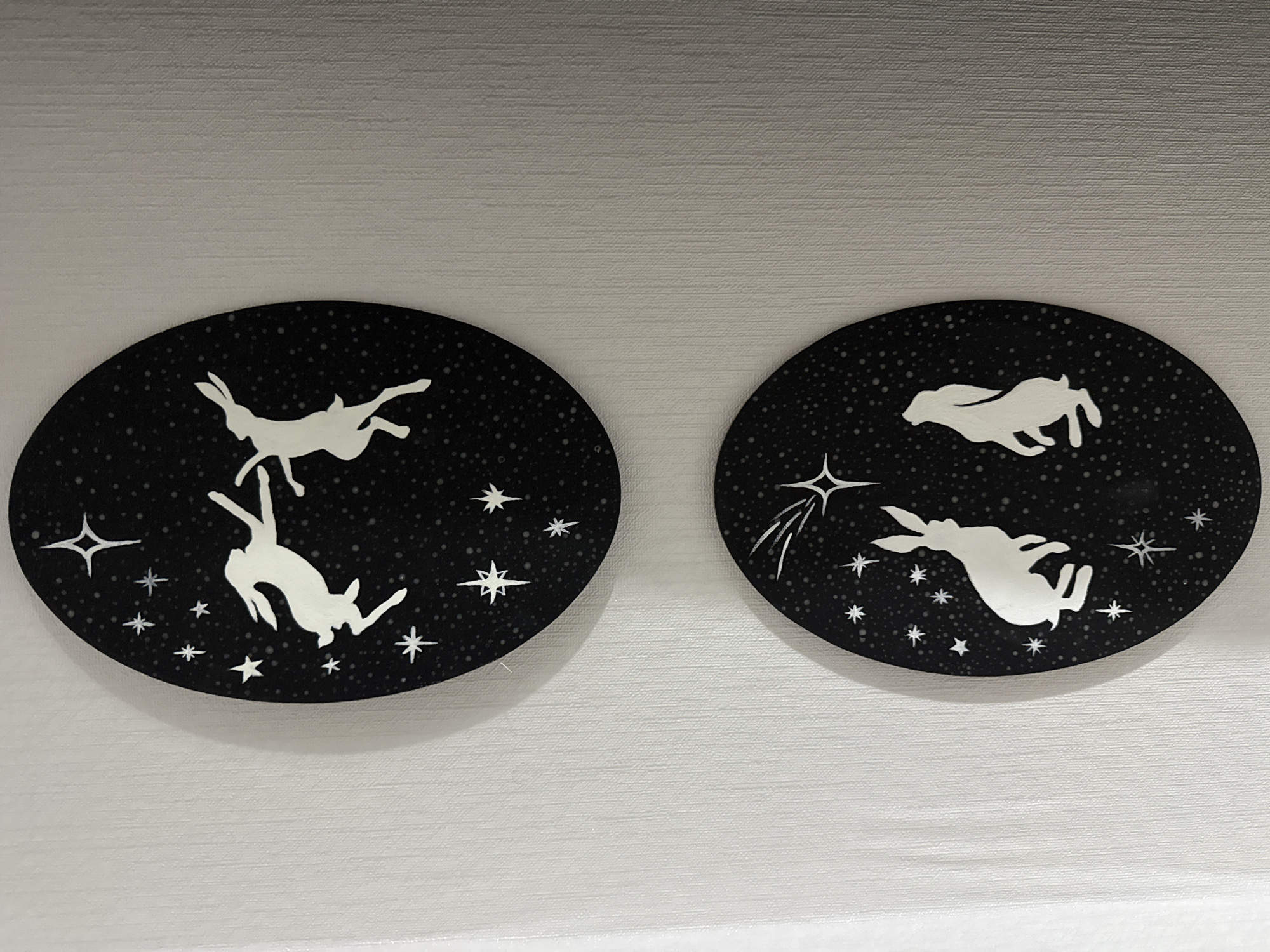 The scene, located in Vniissok, Russia, displays two oval-shaped art panels mounted vertically against a light-colored, vertically textured wall. Each panel features white silhouettes of hares and stars on a dark, speckled background.
The upper panel shows two white hares in dynamic poses against the dark, stippled expanse. The hare on the left is depicted mid-leap, facing right with its front paws extended. The hare on the right is also airborne, reaching its left foreleg towards the left hare's outstretched paw, suggesting an interactive dance. Several white, stylized stars, including one prominent four-pointed star, are scattered across the dark background.
The lower panel features two white hares in contemplative or observing poses. The hare on the left is seated on its haunches, facing right and gazing slightly upwards. The hare on the right stands on its hind legs, looking directly towards the top of the panel. The dark, stippled background includes multiple white, stylized stars. A central star at the top of this panel features a radiating tail, suggesting a shooting star.
The wall exhibits a consistent vertical striation texture, indicative of wallpaper or a similar finish, and appears off-white or light grey. Both art panels cast faint, diffused shadows on the wall, indicating they are slightly raised from the surface. The lighting is even, illuminating the wall's texture and the details of the artwork.