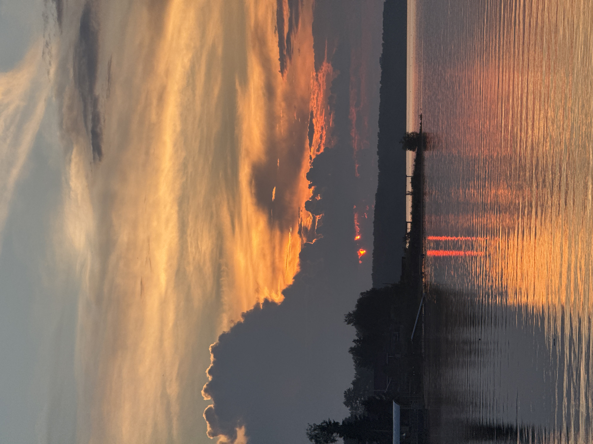 The image depicts a sunset over the Kama River in Chaikovsky, Russia. The sky is filled with vibrant orange and yellow hues, with dark clouds partially obscuring the sun, creating a dramatic effect. The sun's reflection on the rippling water is a prominent feature, casting a warm glow across the surface. Silhouetted against the bright sky are a line of trees and some low-lying buildings, likely residential structures or boathouses, located along the riverbank. A small, dark tree stands on a narrow strip of land extending into the water. The overall scene is one of natural beauty and tranquility, characteristic of a sunset on a body of water. There are no people or animals visible in the image.
