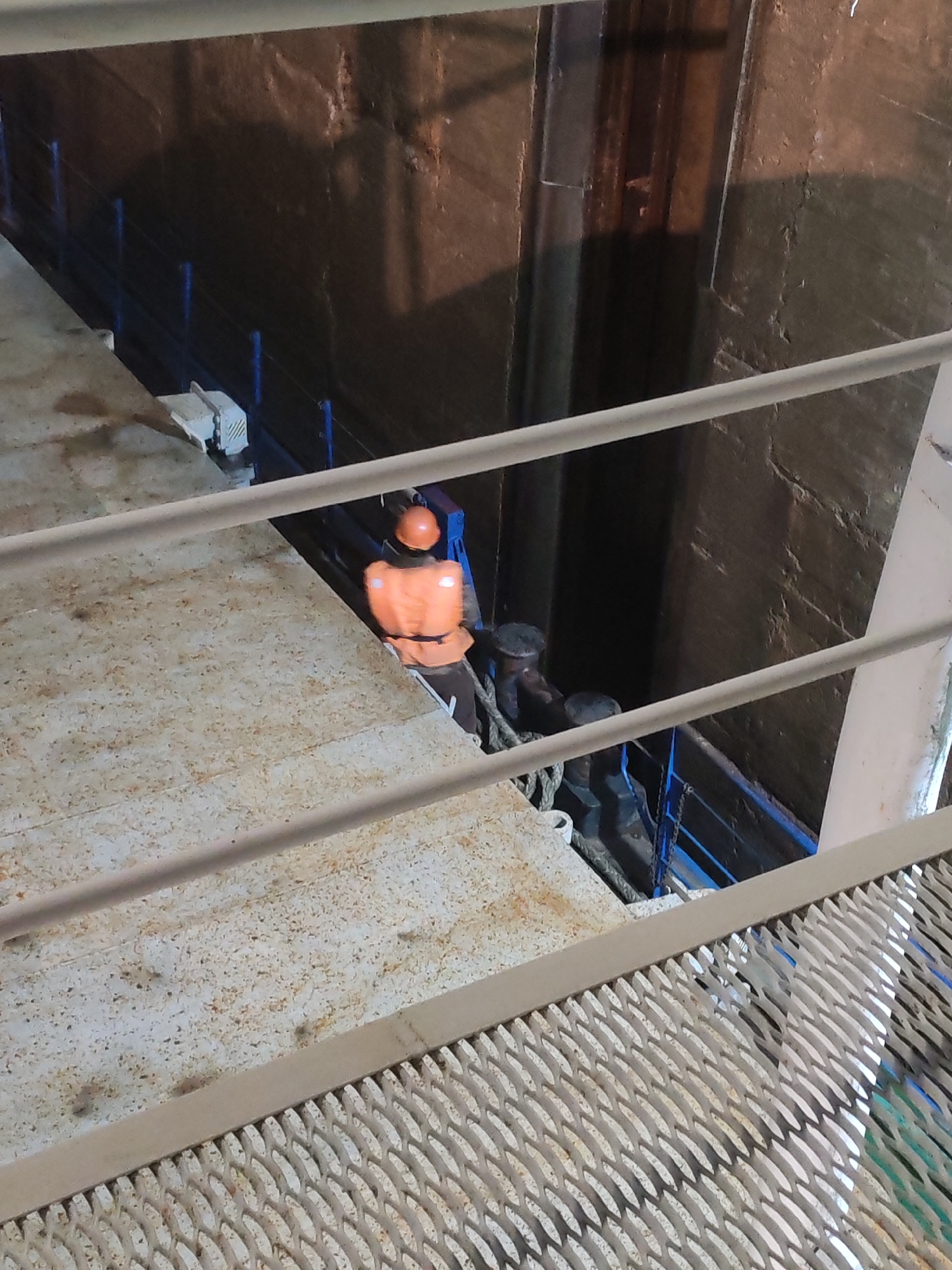 An interior photograph depicts an environment of metallic surfaces and structures, likely a bridge or a dock, with a partial view obstructed by metallic handrails. An individual wearing an orange life vest and a helmet is located on a lower platform. They are standing near bollards, with mooring ropes wrapped around them. To the side of the person, there's a deep, shadowed recess possibly a wall of a lock or waterway. The location is tagged as "Unknown, Unknown."