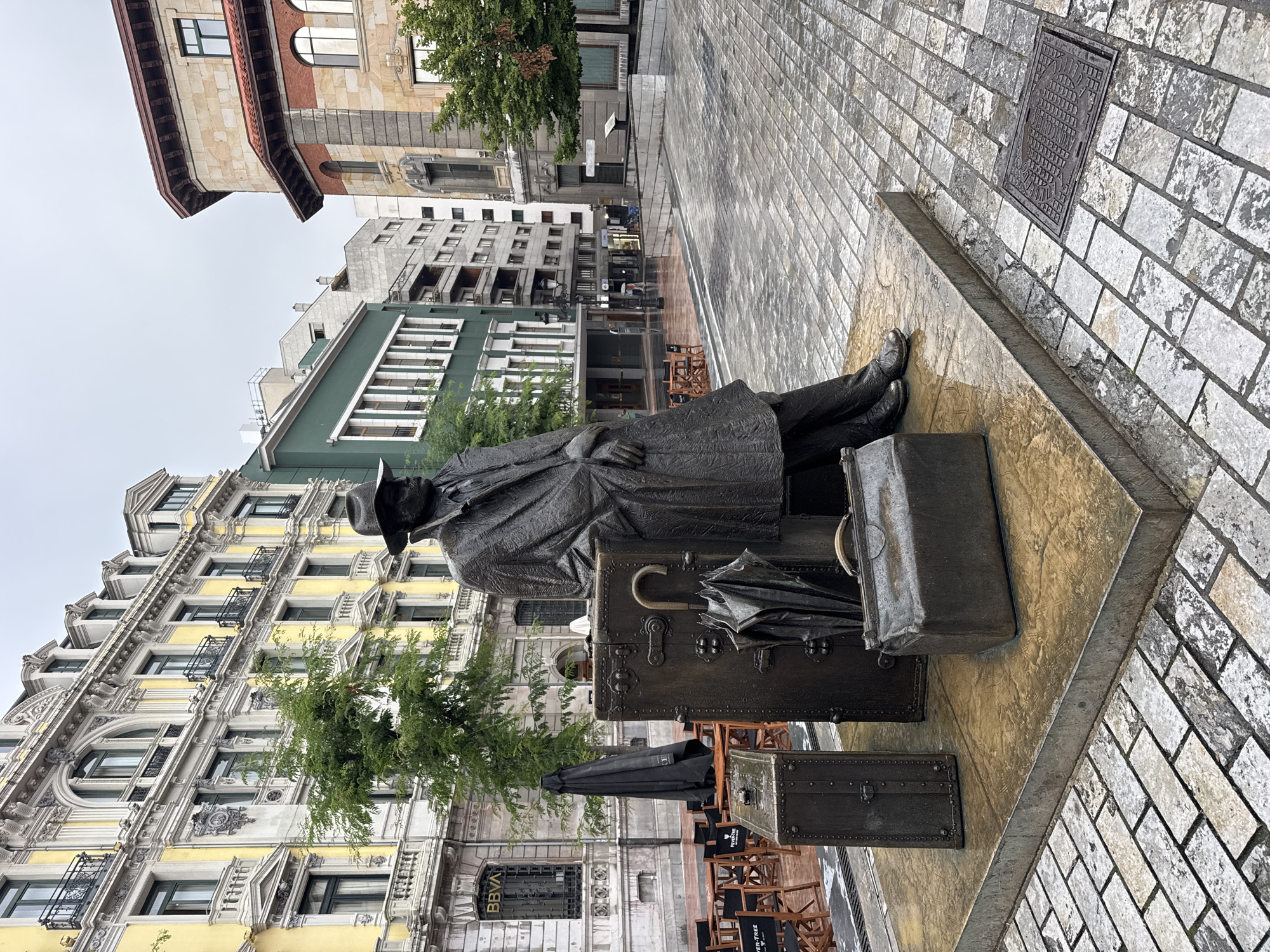 This is a medium shot, eye-level, outdoor, full-color photograph of a bronze statue of a man dressed in a trench coat and fedora, seated with luggage in a European city square. The statue is the central focus, positioned on a raised platform on the right side of the frame. The man is leaning against a large, dark trunk, with a smaller trunk to his left and a briefcase and an open umbrella in front of him. The luggage appears weathered and metallic, with visible latches and hardware. The statue's pose is relaxed, with his right leg crossed over his left.
The setting is an urban plaza with cobblestone paving that appears wet, suggesting recent rain. The architecture in the background is predominantly European in style, featuring multi-story buildings with ornate facades. To the left of the statue, a bright yellow building with neoclassical elements dominates the background, featuring numerous windows and balconies. Adjacent to it, a bright green building with a more modern design is visible. Further in the background, other buildings of varying architectural styles and heights are present, with some trees and streetlights interspersed. Outdoor seating areas with tables and chairs, likely for a cafe or restaurant, are visible on the left side of the frame, in front of the yellow building. A black umbrella is deployed and closed, resting against the yellow building. The sky is overcast and a pale grey. The overall atmosphere is subdued, possibly due to the weather.
The statue is located in the Plaza de la Escandalera in Oviedo, Spain.