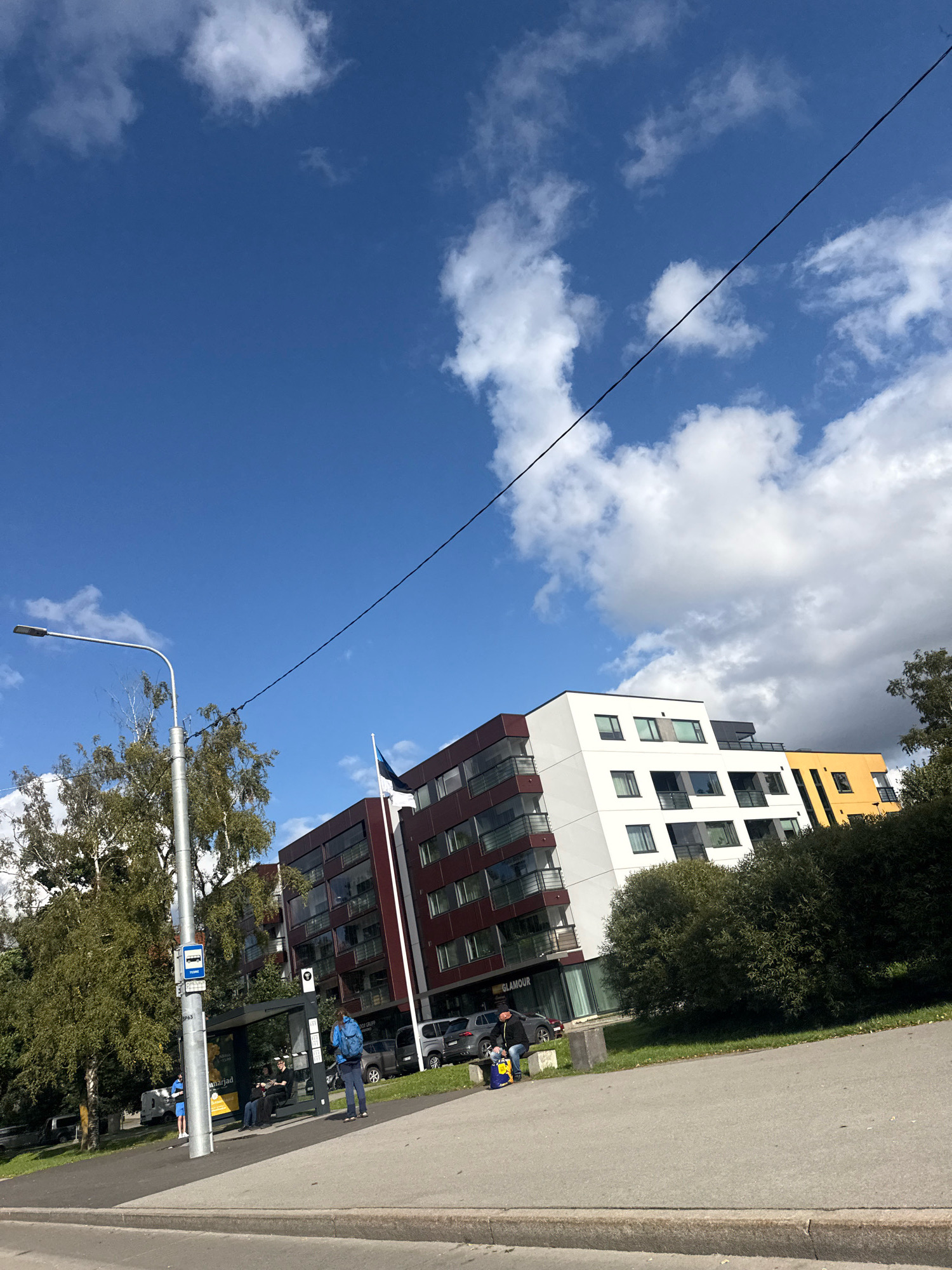 The image captures a street scene in Tallinn, Estonia, on a partly cloudy day. A modern apartment building with sections of dark red, white, and yellow facades dominates the background, featuring numerous windows and balconies. To the left of the apartment building, an Estonian flag flies from a flagpole. In the foreground, a bus stop shelter is visible, with several people waiting. A person wearing a blue backpack stands near the shelter, while others are seated. Adjacent to the bus stop, a tall lamp post stands with a street light and overhead wires stretching across the frame. Lush green trees and bushes line the area behind the bus stop and in front of the buildings. A paved road and sidewalk are in the foreground, with a curb separating them from the street. Several cars are parked near the buildings and on the street. The sky is a bright blue, dotted with white, fluffy clouds.