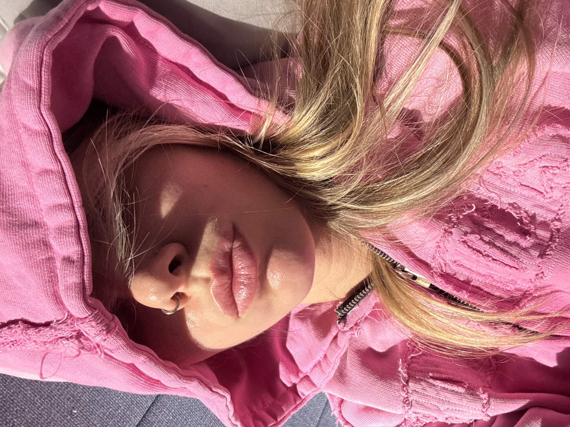 In an indoor close-up, a person is lying down, wearing a pink hooded jacket with frayed edges. Their face is partially obscured by the hood, with a silver nose ring visible. Their blonde hair is spread across the pink fabric. Sunlight casts shadows on their face and hair. The background is indistinct, suggesting an indoor setting.