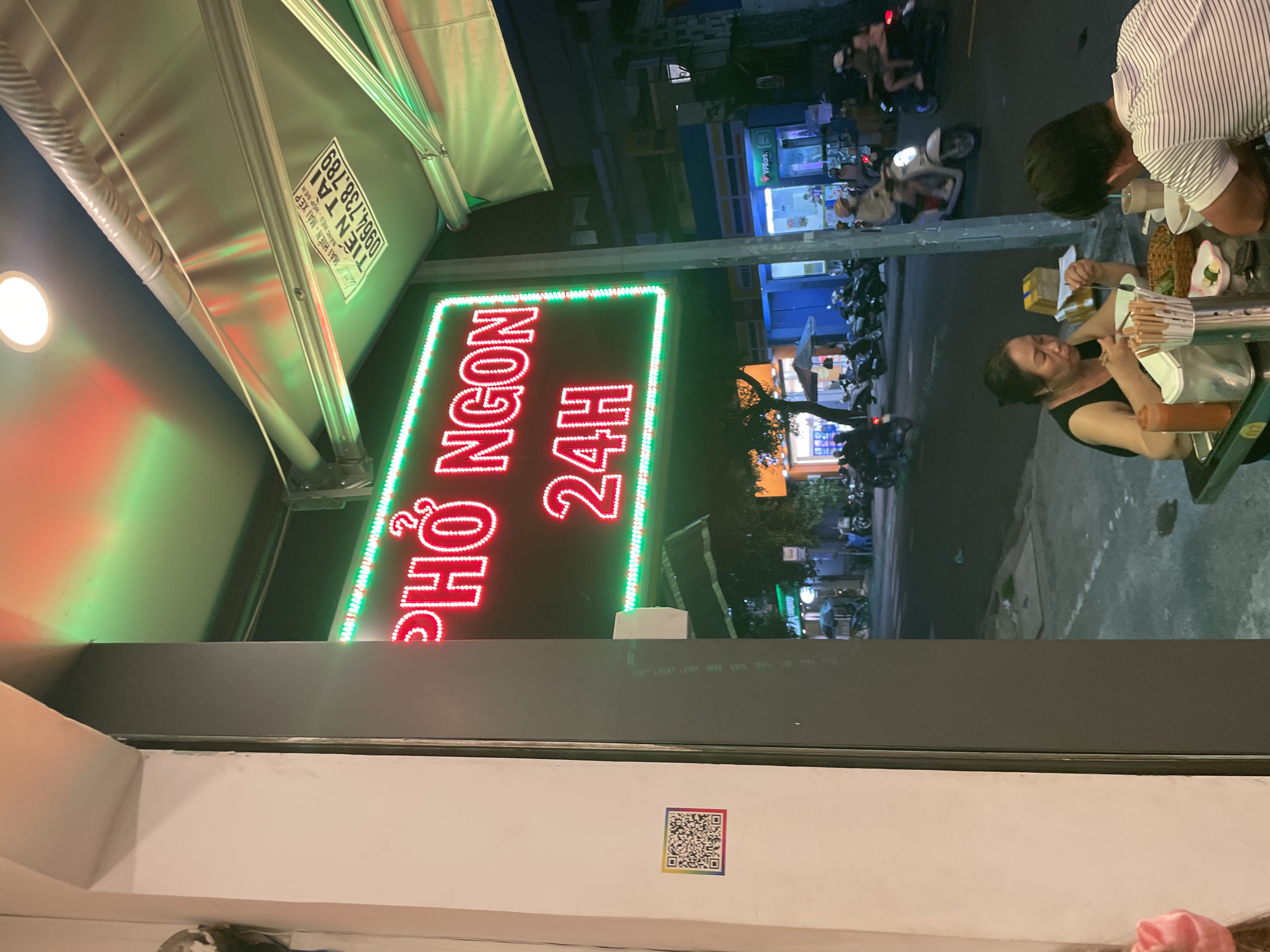 This is a nighttime, medium shot of a street scene in Thủ Đức, Vietnam, focused on a restaurant with a prominent neon sign. The sign, illuminated in red and green, reads "PHỞ NGON 24H" indicating that pho is served 24 hours a day. To the left of the sign, a small, colorful QR code is affixed to a pale wall.
In the foreground, two individuals are seated at a table, appearing to be eating. A woman in a black tank top is on the left, actively eating with chopsticks. A man in a striped shirt is on the right, facing away from the camera. Various dining implements, including chopsticks, bowls, and a bottle of sauce, are present on the table.
The background reveals a busy street with parked motorcycles and several people. Buildings with illuminated storefronts line the street, suggesting a commercial area. The overall impression is a lively urban environment during evening hours.