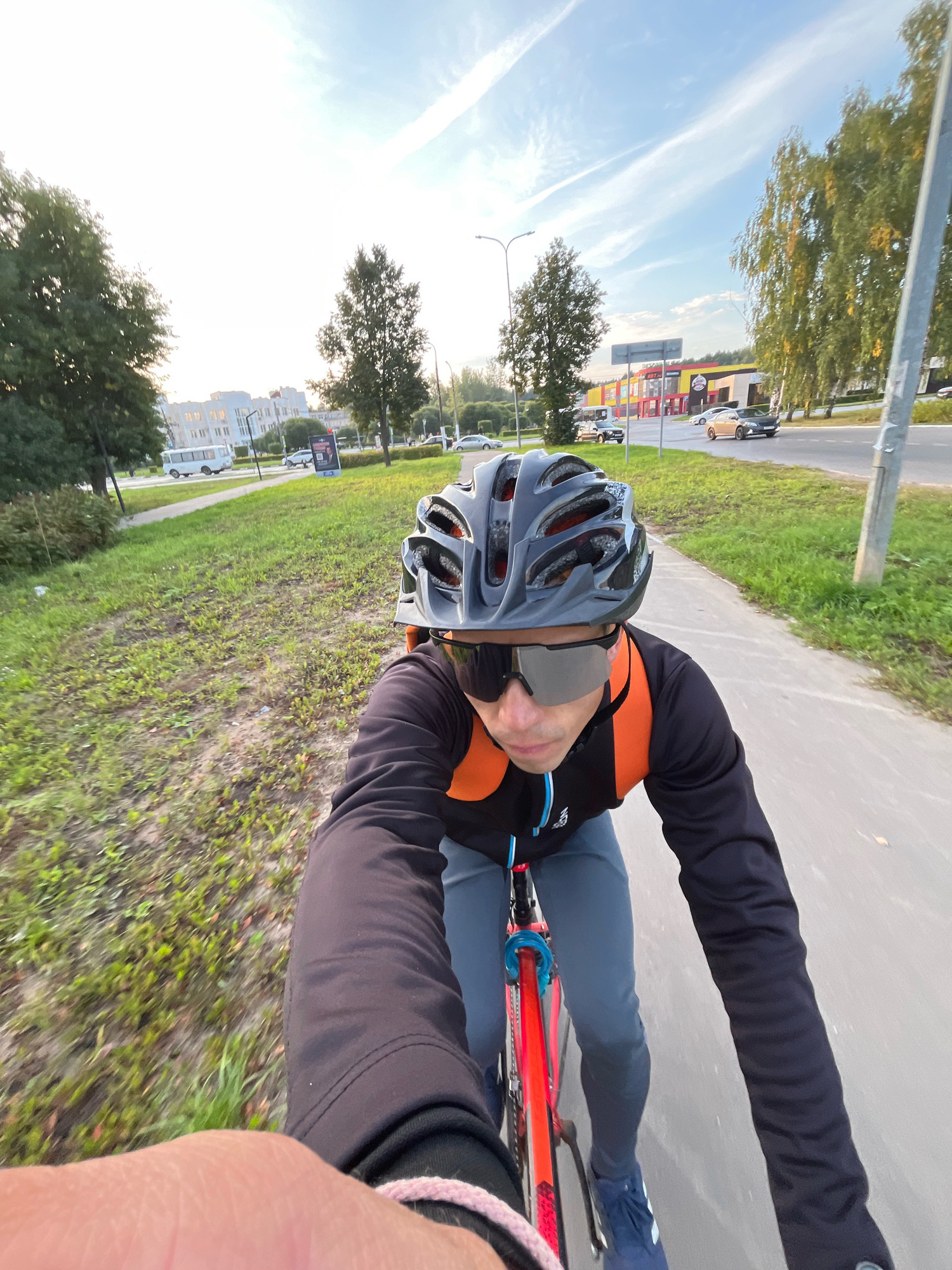 In Chaikovsky, Russia, on a paved path next to a grassy area and a road, a person rides a bicycle. The cyclist, wearing a black long-sleeved shirt, blue pants, blue athletic shoes, a grey helmet, and sunglasses, is leaning forward. An orange backpack is visible on their back. The road has several cars, including a white minibus, a black SUV, and a gold sedan. On the right side of the road, a streetlamp stands tall, and birch trees with yellowing leaves line the roadside. In the distance, several buildings are visible, including a brightly colored commercial building with signage. The sky is blue with streaks of white clouds, and the sun is low, casting long shadows.