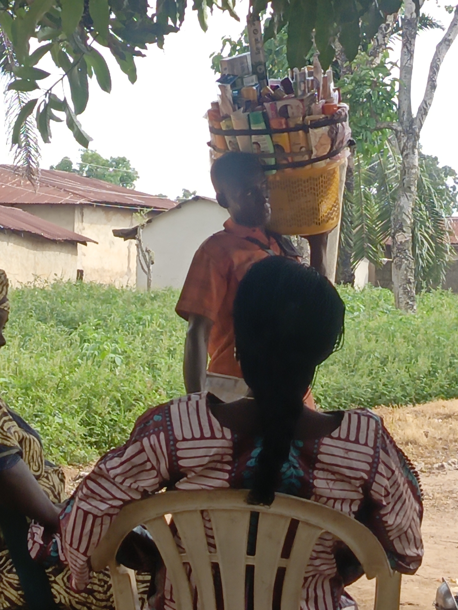 The image shows a scene in Unguwar Zaria, Nigeria. A person wearing an orange shirt carries a large yellow basket on their head, filled with various items. Two people are sitting in the foreground, with the back of one person in a patterned shirt prominently visible. The surrounding environment consists of green vegetation, trees, and simple buildings with rusty roofs. A dirt road is also visible.