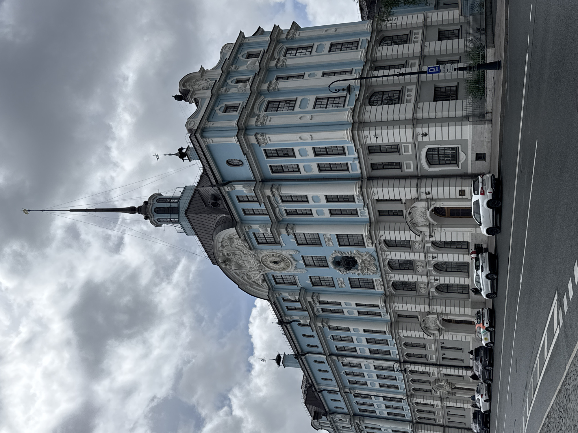 The image captures the Smolny Institute, a historical building in Saint Petersburg, Russia, from a ground-level perspective, rotated 90 degrees clockwise. The architecture features a predominantly blue and white façade, with multiple stories, numerous windows, ornate detailing, and a prominent spire extending upwards into the cloudy sky. Parked vehicles line the street adjacent to the building. The sky is overcast with predominantly gray clouds.