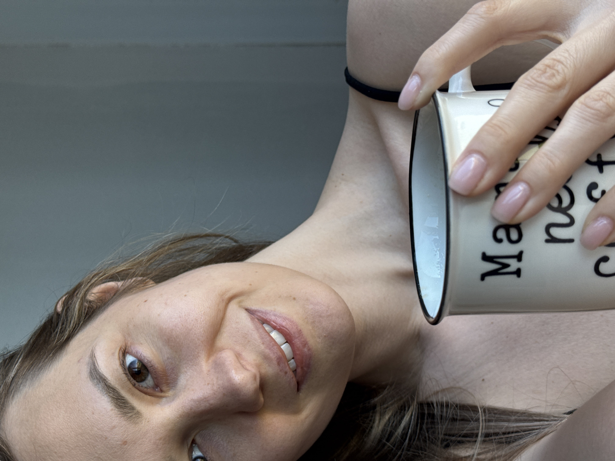 A woman with fair skin and brown hair is holding a mug with white liquid in it near her neck. She is smiling and looking into the camera. The mug is white with black letters. A thin black choker is around her neck. The background appears to be a plain, light gray surface. There is no indication of the city of Chaikovsky, Russia, in the image.