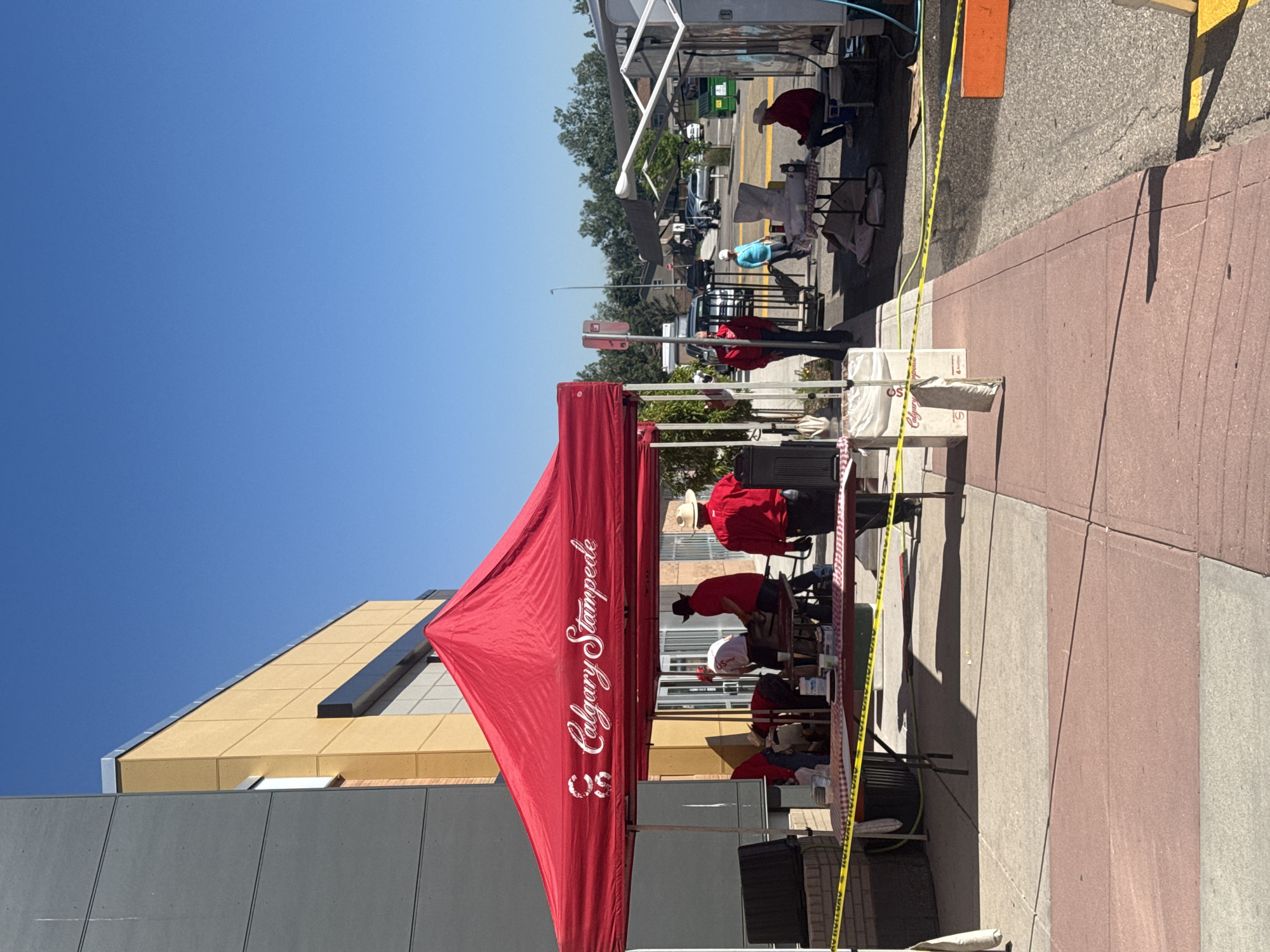 In Calgary, Alberta, Canada, under a clear blue sky, a Calgary Stampede event is underway outdoors. A large red tent with the Calgary Stampede logo is erected, providing shade for several individuals. People wearing red shirts and cowboy hats are actively engaged in various tasks beneath the tent, some appearing to be preparing food or interacting with supplies. A checkered tablecloth covers a table within the tent, and yellow caution tape cordons off the immediate area.
Beyond the tent, the scene continues with more activity. Another tent, possibly a food or merchandise booth, is visible further down the sidewalk. Several people are present, including one individual seated and another walking. In the background, a street with parked cars and buildings can be seen, suggesting an urban or suburban setting. The ground is paved with concrete and red bricks.