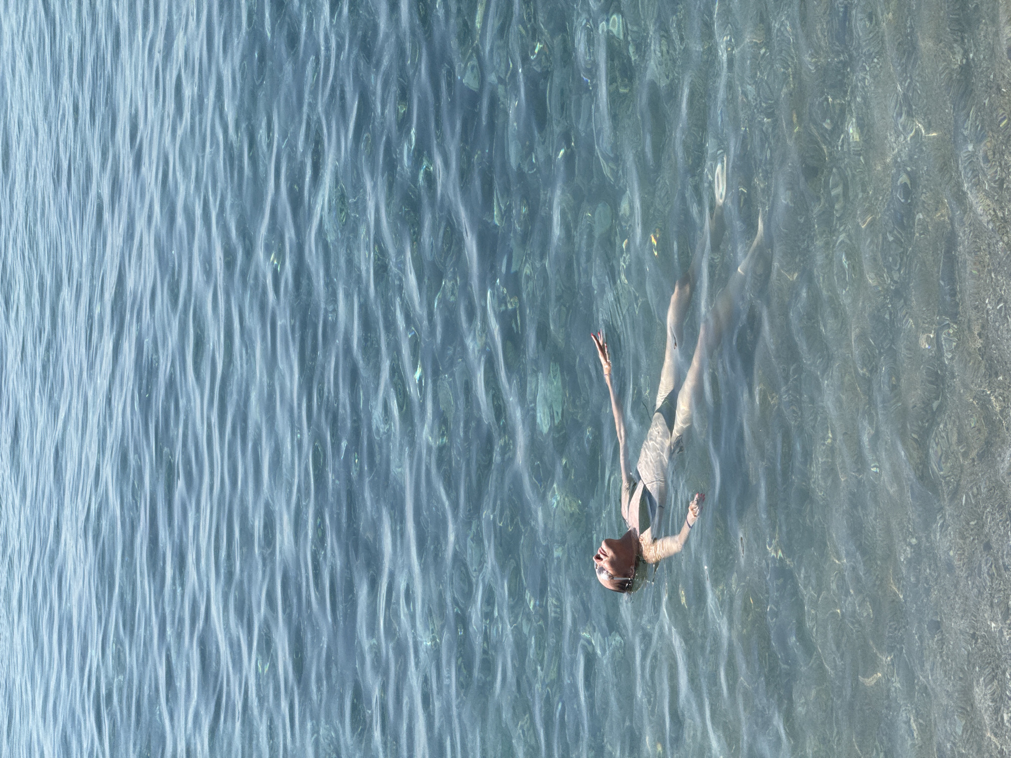 A person is floating on their back in clear, shallow water. The water has visible ripples and the sandy or pebbly bottom is visible beneath the surface. The person is wearing sunglasses and a dark-colored swimsuit. Their arms are extended out to their sides, and their head is tilted back, with their eyes closed. The lighting suggests it is a sunny day. The location is not specified in the image to be the city of Abang, Indonesia.