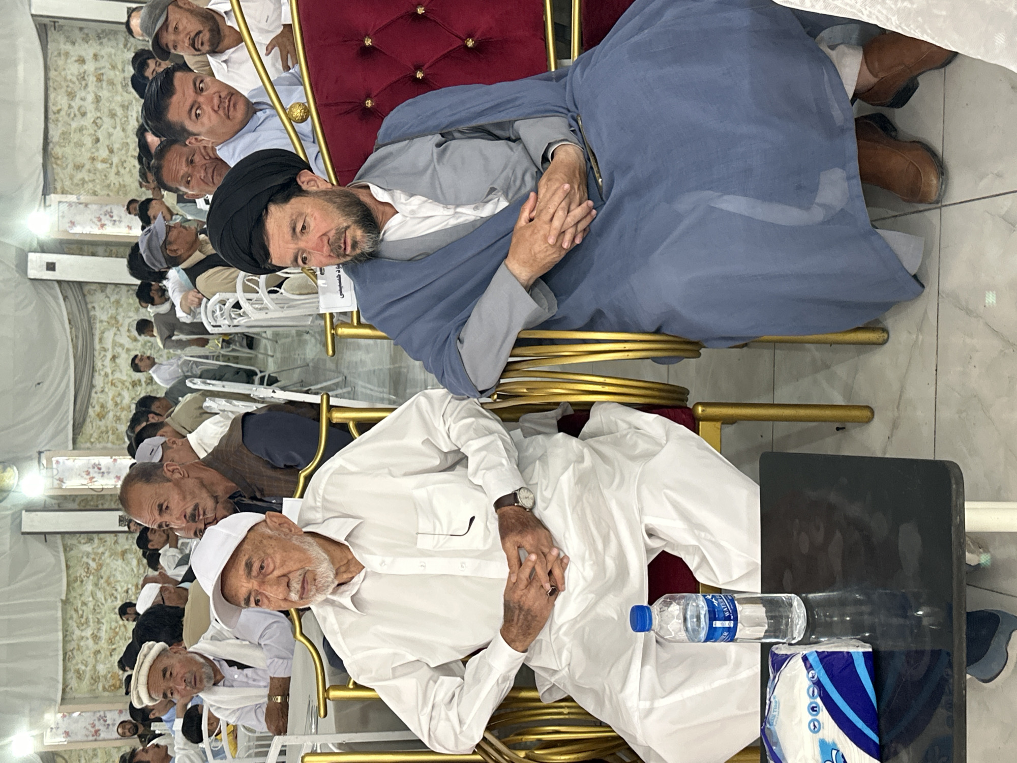 The image shows a gathering of men indoors in an unknown location in the city of Unknown, Unknown. The room has patterned walls and tile floors. The men are mostly seated on gold-colored chairs. A senior man in the foreground wears a white tunic and cap, with a watch on his wrist. A water bottle and tissue box sit on a black table next to him. Another man with a black turban and a light blue shawl sits in the background, resting against a red velvet cushion. Other men, dressed in various white and light-colored garments, are seated in rows behind them.