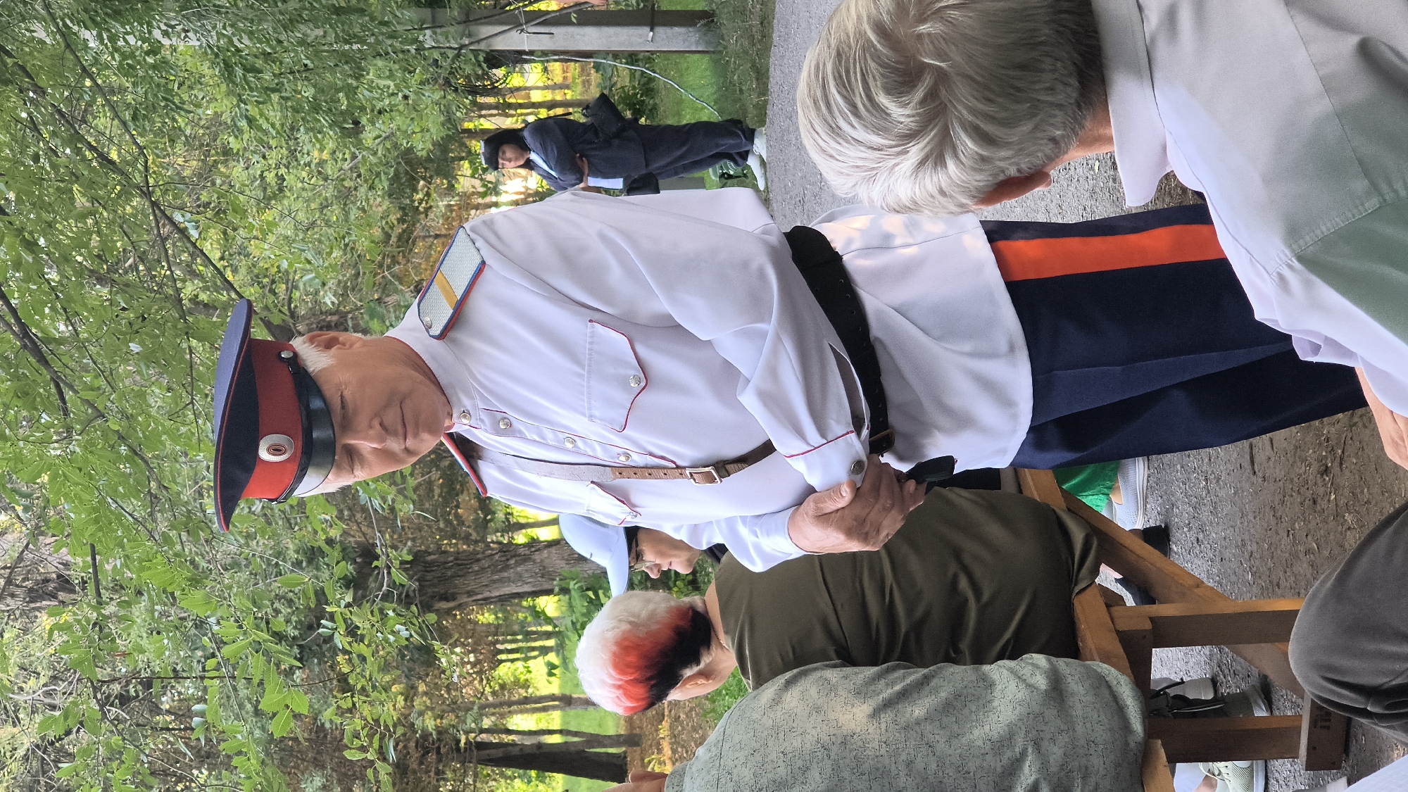 An elderly Caucasian man in a white uniform with red trim, a dark belt, and a traditional Cossack hat stands in a park setting. He has gray hair and is looking down with his hands clasped. Behind him, other people are visible, some seated on a wooden bench, and a woman stands in the background. The environment consists of trees and a gravel path. The scene appears to be outdoors in a public space.