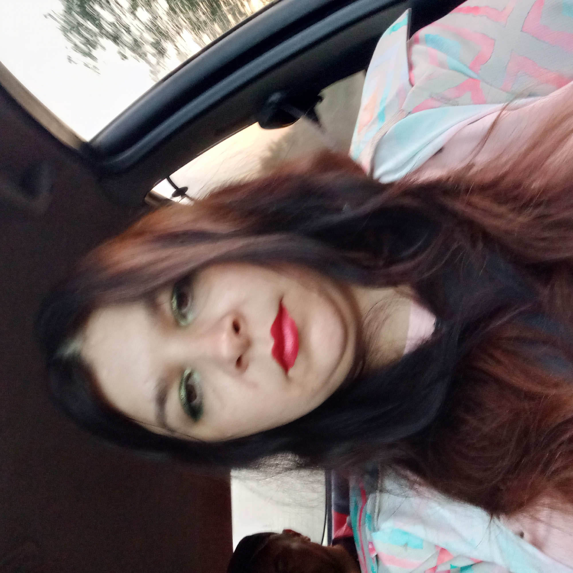 The image shows a close-up selfie taken from inside a car, with the driver likely being the woman in the foreground. She has dark hair and is wearing makeup, including green eyeshadow and red lipstick. She is wearing a pink shirt and a light-colored jacket with a colorful geometric pattern. In the background, a person wearing a dark cap is visible in the passenger seat. The car appears to be in motion, as suggested by the blurred trees and sky visible through the windows. The location is specified as Klyntsi, Ukraine.