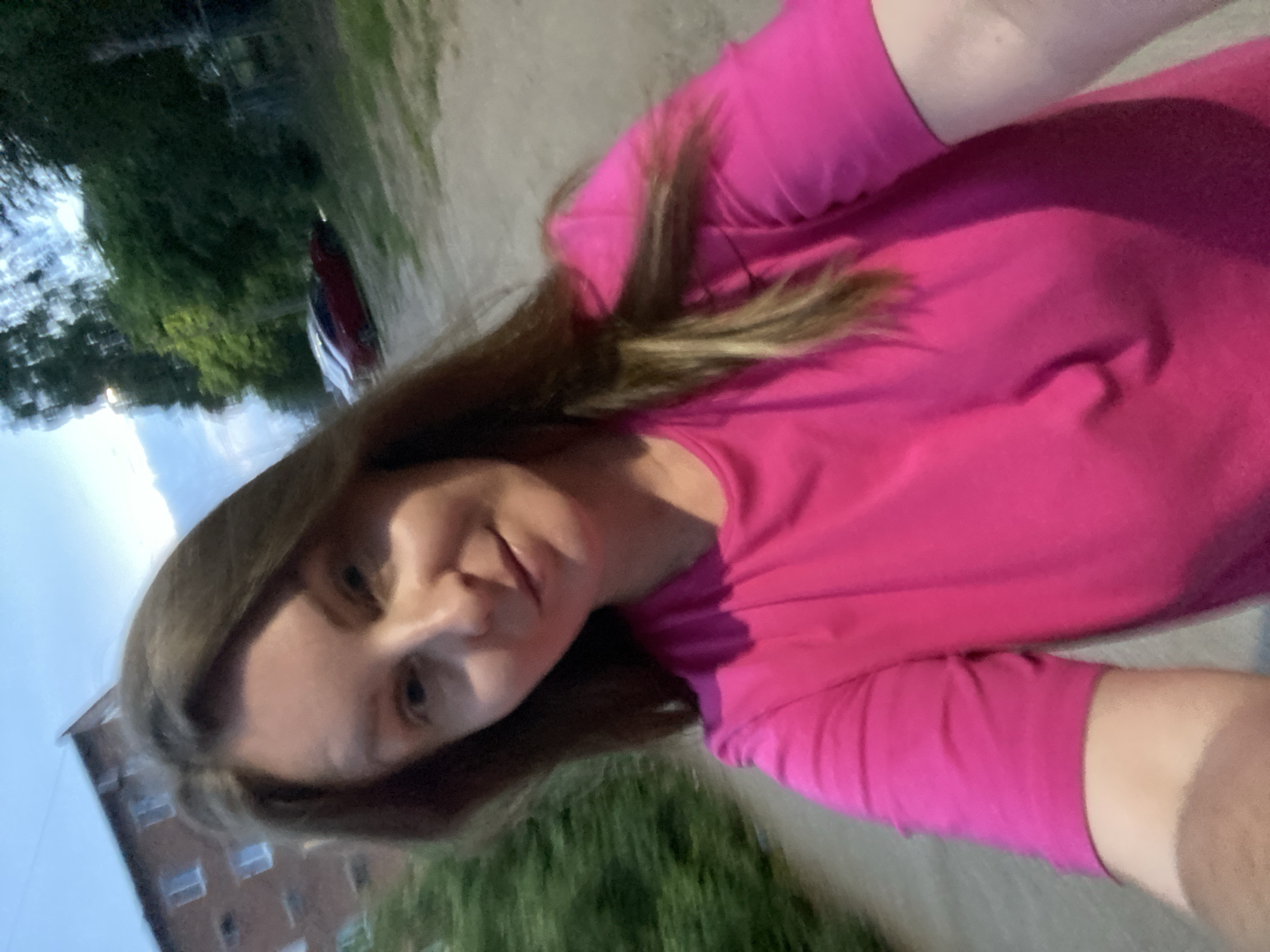 This is a selfie taken outdoors, possibly in Noviy, Russia. The subject is a person with brown hair and light skin, wearing a bright pink t-shirt. They are looking towards the camera with a slight smile.
The background is blurred, but it appears to be a residential area. To the left, there's a building with multiple windows, suggesting an apartment complex. In the distance, there are trees and green foliage, indicating an outdoor setting with some greenery. The ground appears to be a paved or dirt path. The lighting suggests it might be daytime, possibly late afternoon or early evening, as the sky is visible and somewhat bright. There is no visible text in the image. The overall mood appears casual and relaxed.