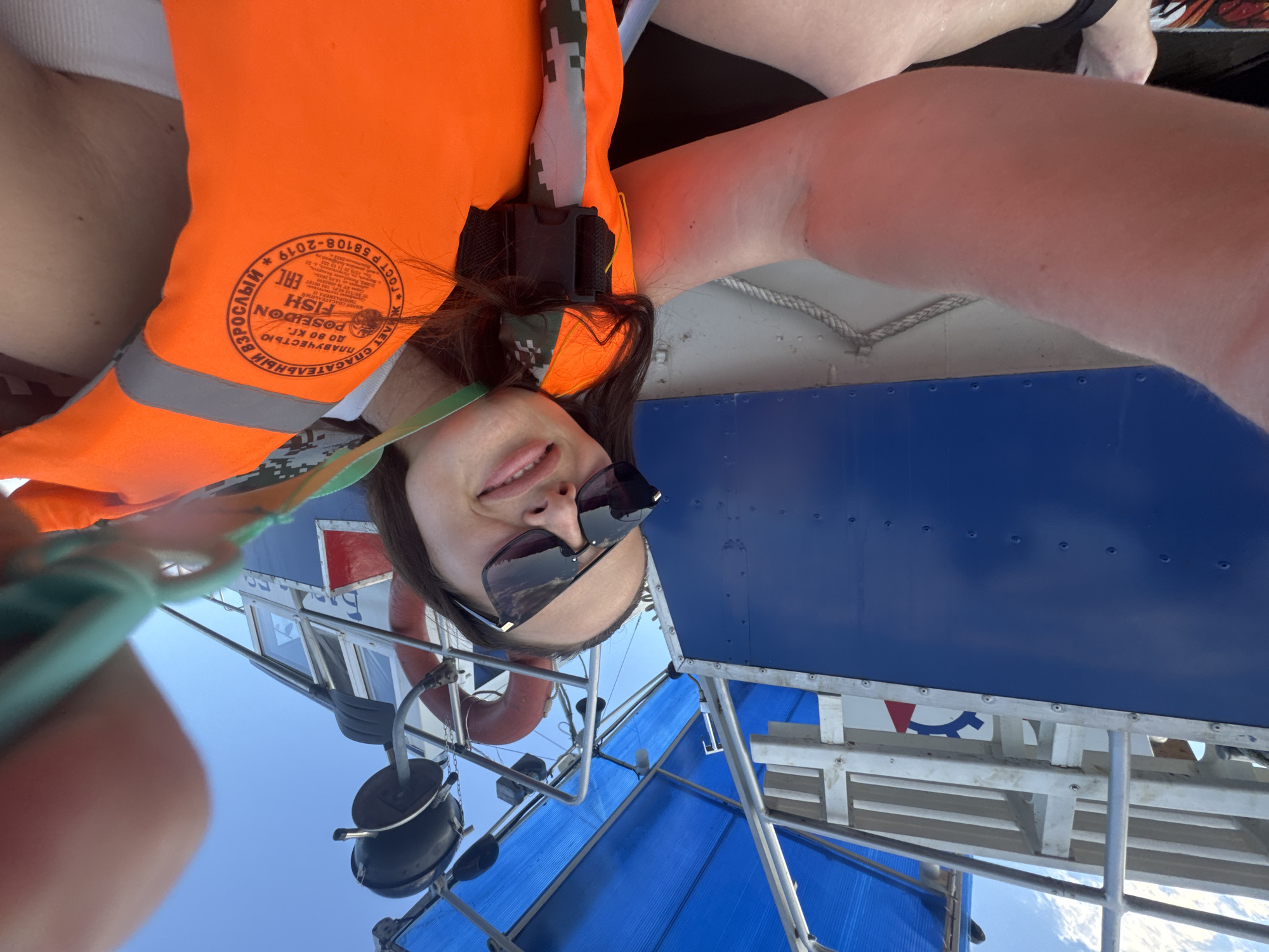 A young woman with long, dark hair and sunglasses smiles at the camera while on a boat. She is wearing an orange life jacket over a white shirt. The boat has a blue hull and a blue canopy. The background shows a clear blue sky. The image is taken from a low angle, looking up at the woman and the boat. The location is identified as Chaikovsky, Russia.