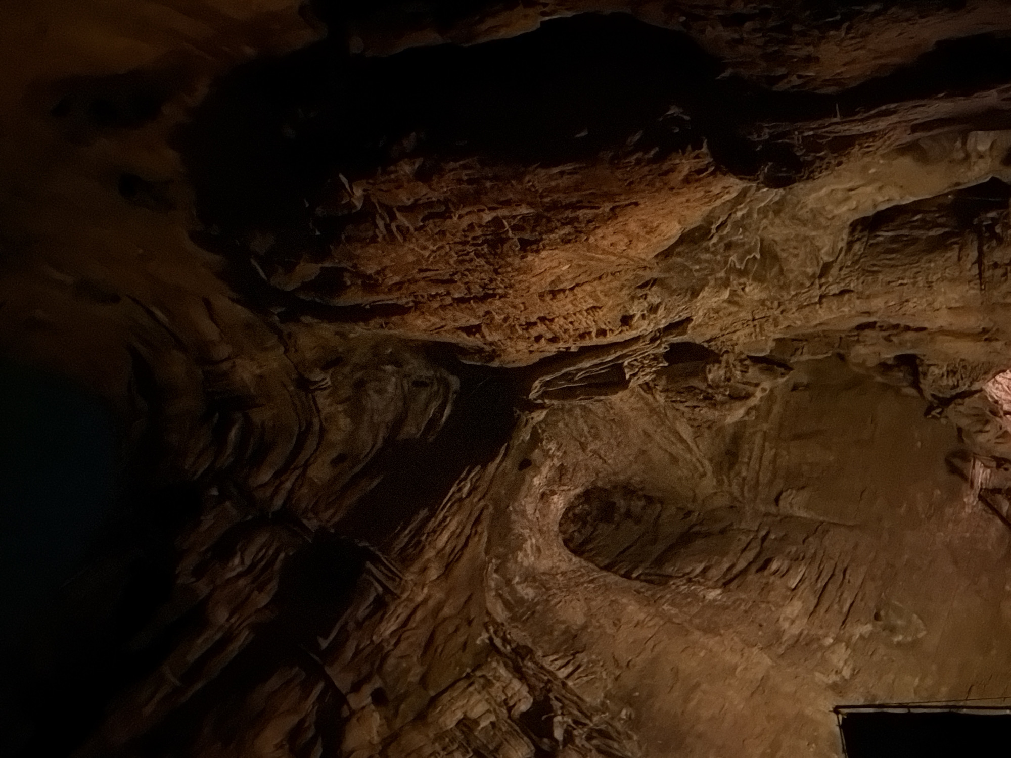 The image shows the interior of a cave with rough, textured rock formations illuminated by artificial lighting. The rock walls display layers and striations, with areas of deep shadow contrasting with patches of warm orange and brown light. In the lower left corner, a dark, rectangular object, possibly an entrance or a piece of equipment, is partially visible. The overall environment is dark, cavernous, and appears to be a natural geological formation. There are no people or animals visible in the image. The image does not provide specific visual cues to confirm its location in Guttapadu, India.