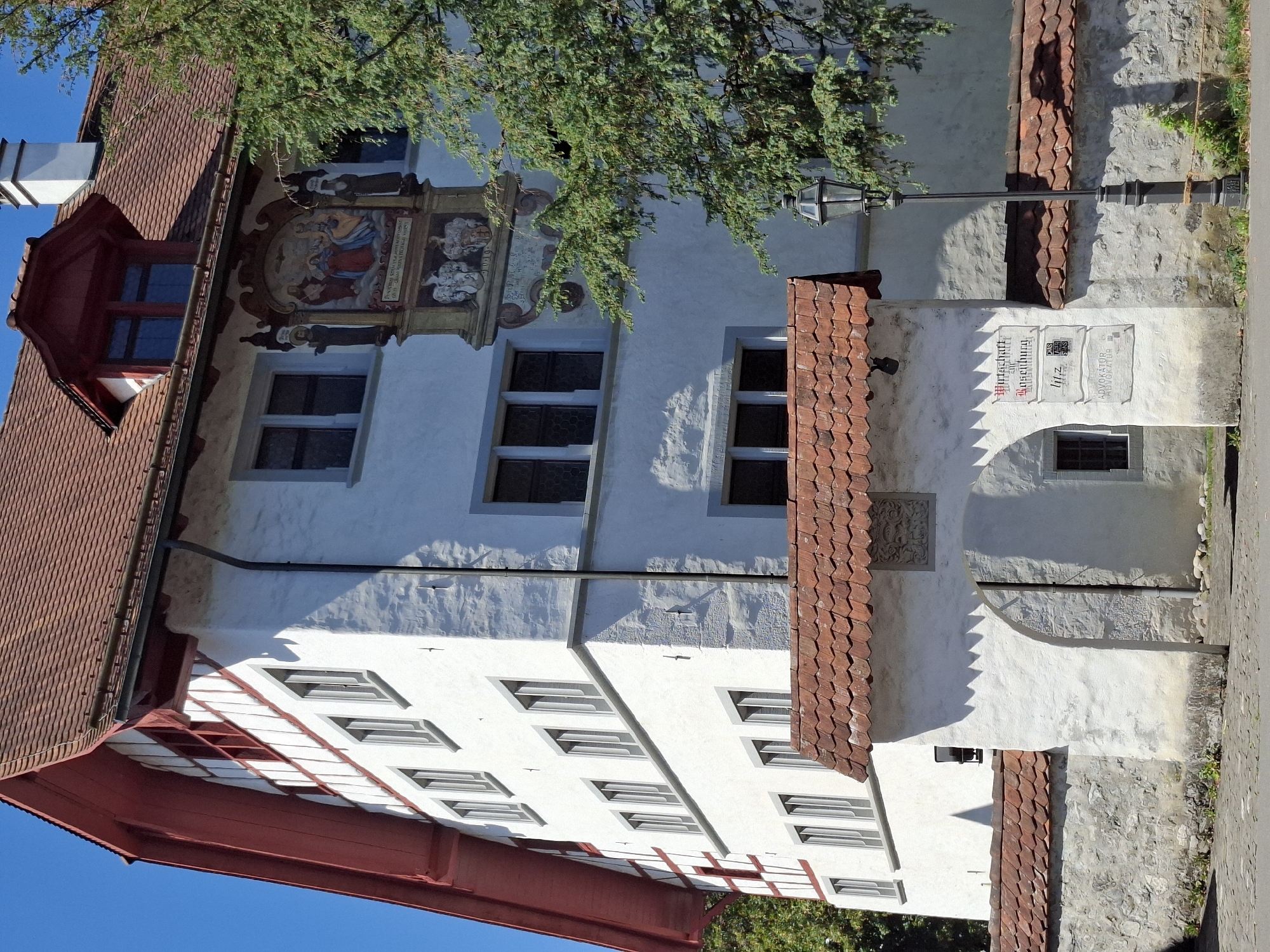 The image shows the historic building of the "Kultur und Kongresszentrum Oberdorf" in Stans, Switzerland. The building features a traditional Swiss architectural style with a white plastered facade, red-tiled roofs, and dormer windows. A large fresco depicting a religious scene is mounted on the facade. An arched entrance leads to a courtyard. The scene is bathed in sunlight, casting shadows on the building and surrounding stone walls. A street lamp stands to the right of the entrance. The location is identified by signs on the wall of the arched entrance, which include "Wirtschaft zum Winkelburg" and "ADVOKATUR".