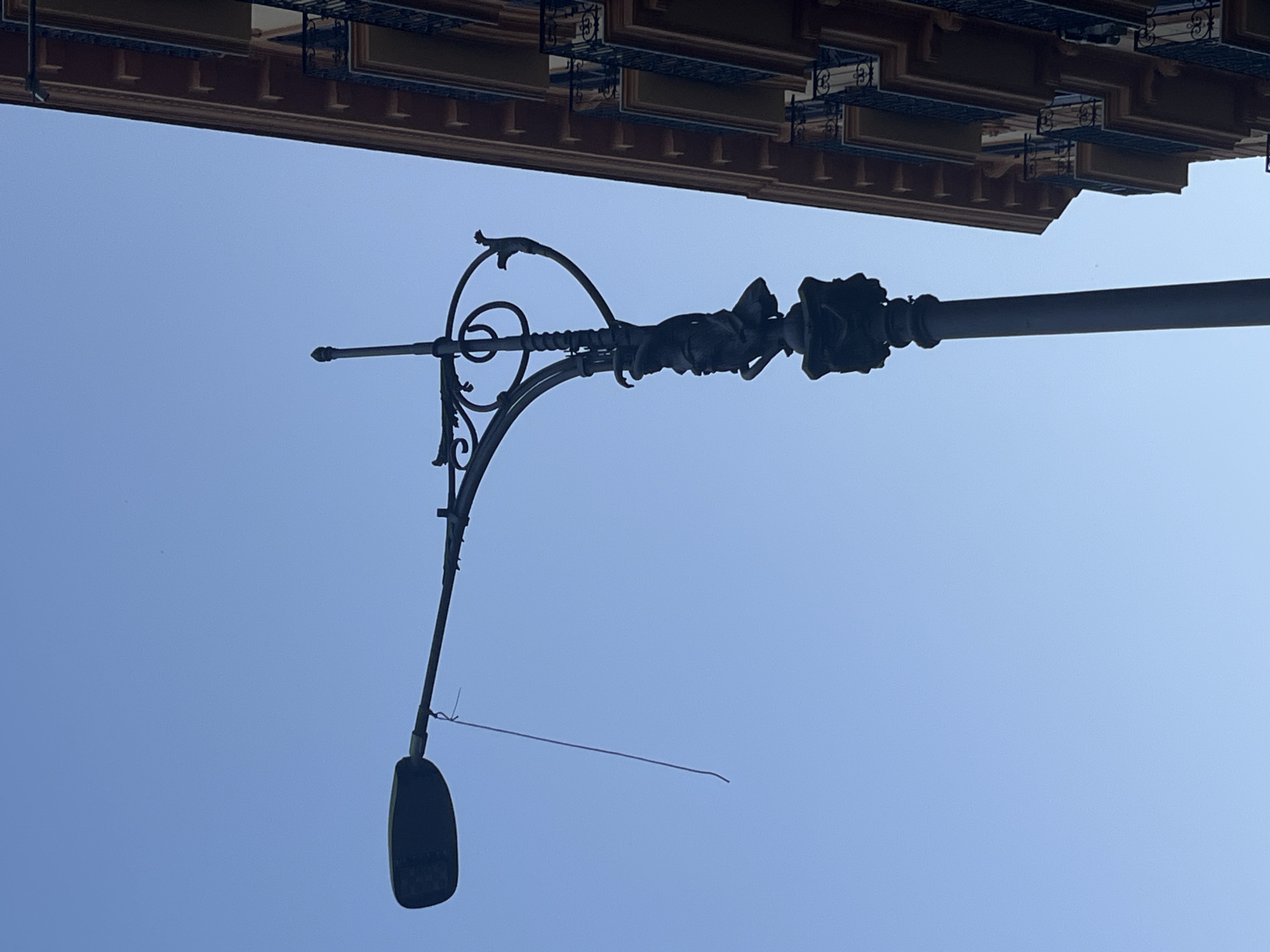 This image depicts a lamppost in Naples, Italy, against a clear blue sky. The lamppost is ornate, with decorative scrollwork and a sculpted figure. A thin wire or string hangs from the arm of the lamppost, dangling in the air. To the right of the lamppost, a portion of a building with balconies and intricate architectural details is visible. The scene appears to be in an urban setting, likely a street or piazza in Naples.