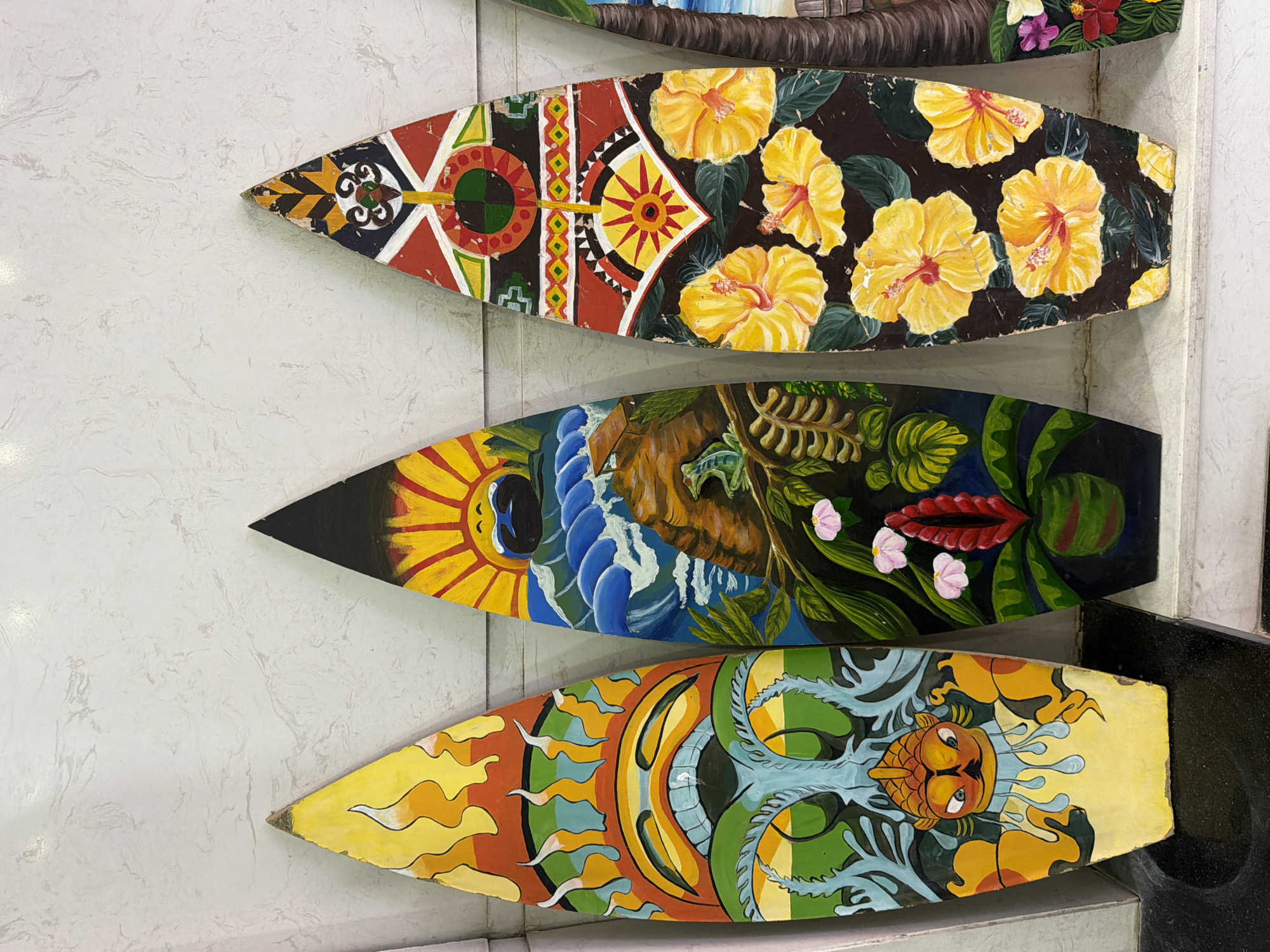Four decorative surfboards are displayed vertically against a textured, off-white wall. The surfboards feature vibrant, hand-painted designs. The leftmost surfboard has a yellow base with abstract orange and yellow flame-like motifs at the top, transitioning to a green design resembling foliage and a stylized fish-like face with blue eyes and orange accents at the bottom. The second surfboard from the left depicts a tropical scene with a smiling sun at the top, blue waves crashing against a rocky outcrop, and lush green foliage with small pink flowers and a red bloom at the bottom. The third surfboard from the left has a dark background adorned with large, yellow hibiscus flowers with red centers and green leaves, interspersed with colorful geometric patterns at the top. The fourth object, partially visible on the right, appears to be another decorative item, possibly a surfboard, with a painted palm tree and floral motifs. The surfboards are positioned side-by-side on a dark surface, likely a shelf or counter, with a white ledge running beneath them. The scene is lit by an overhead light source, creating soft shadows. The setting is likely a shop or market stall in Da Nang, Vietnam, given the tropical theme and artisanal nature of the objects.