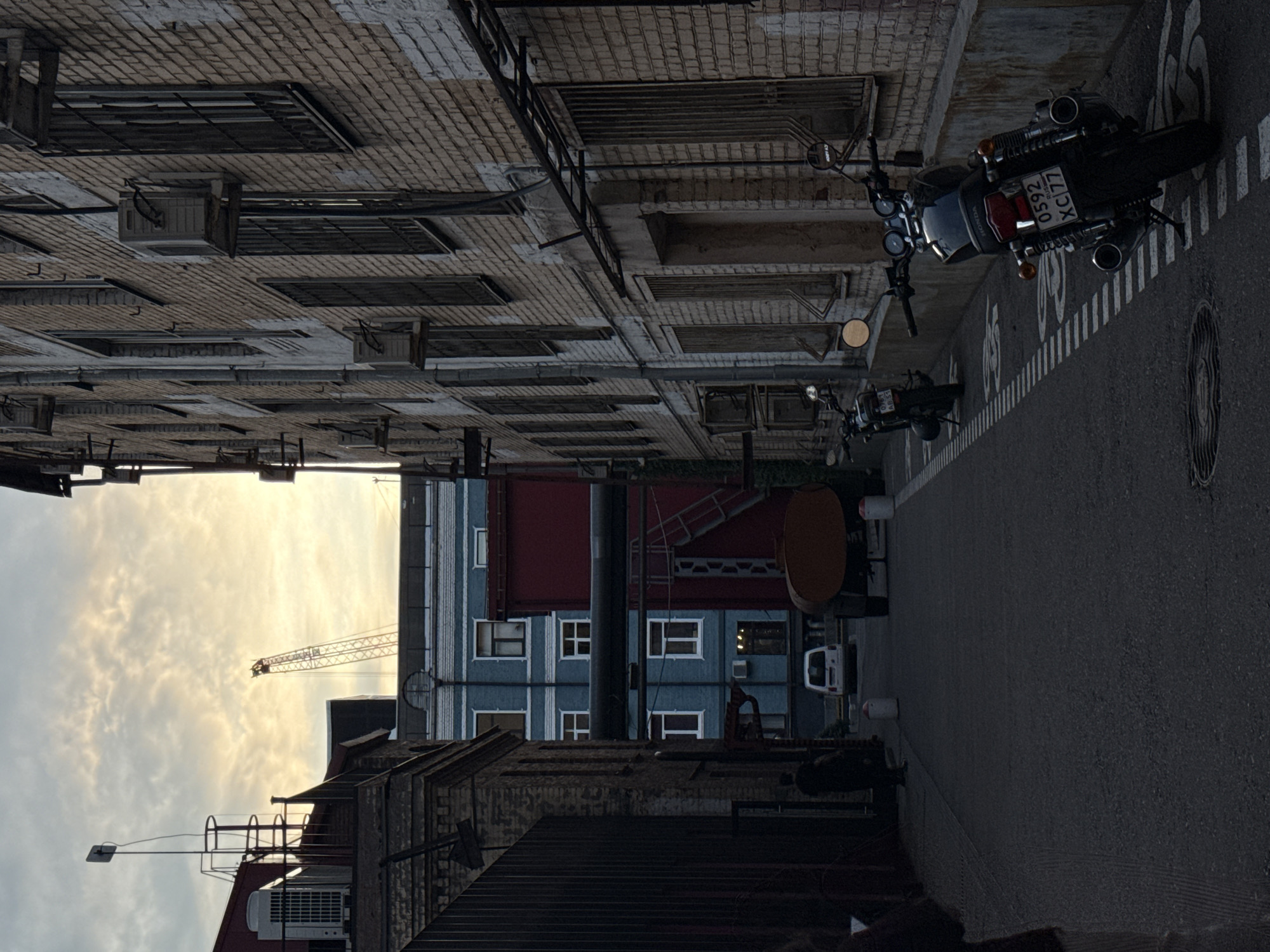 The image depicts an industrial alleyway in Moscow, Russia, at dusk or dawn. On the right, a weathered brick building with multiple windows, some barred, lines the alley. Air conditioning units are mounted on its exterior. Two motorcycles are parked on the left side of the alley, one in the foreground with a Russian license plate "0592 XC77" and another further back. The alley pavement is marked with white lines, possibly indicating a bike lane. In the background, a light blue apartment building with white-trimmed windows stands between two darker industrial structures, one red. A white van is parked in front of the blue building. The sky above is cloudy with a warm glow, and a construction crane is visible in the distance. A few indistinct figures are present in the alley.