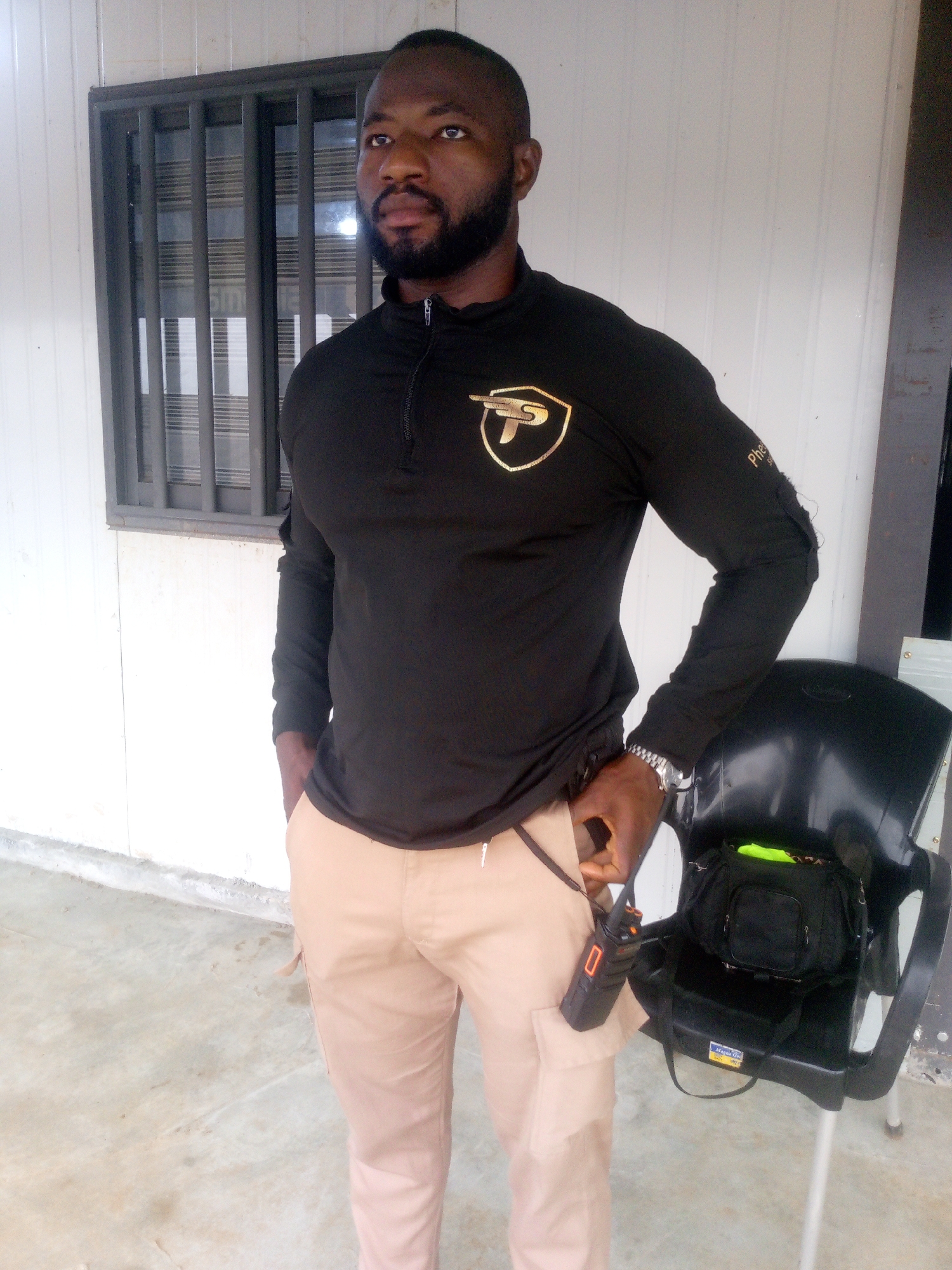 A Black male, appearing to be in his late 30s or early 40s, stands outdoors in front of a white, corrugated metal building with a window that has vertical bars. He is wearing a black, long-sleeved shirt with a gold emblem on the chest and beige cargo pants. A black walkie-talkie is clipped to his belt and hangs from a lanyard around his neck. His left hand is tucked into his pants pocket. A black plastic chair is to his right, with a black bag and a bright green object on it. The ground is concrete. The scene is located in Indo, Nigeria.