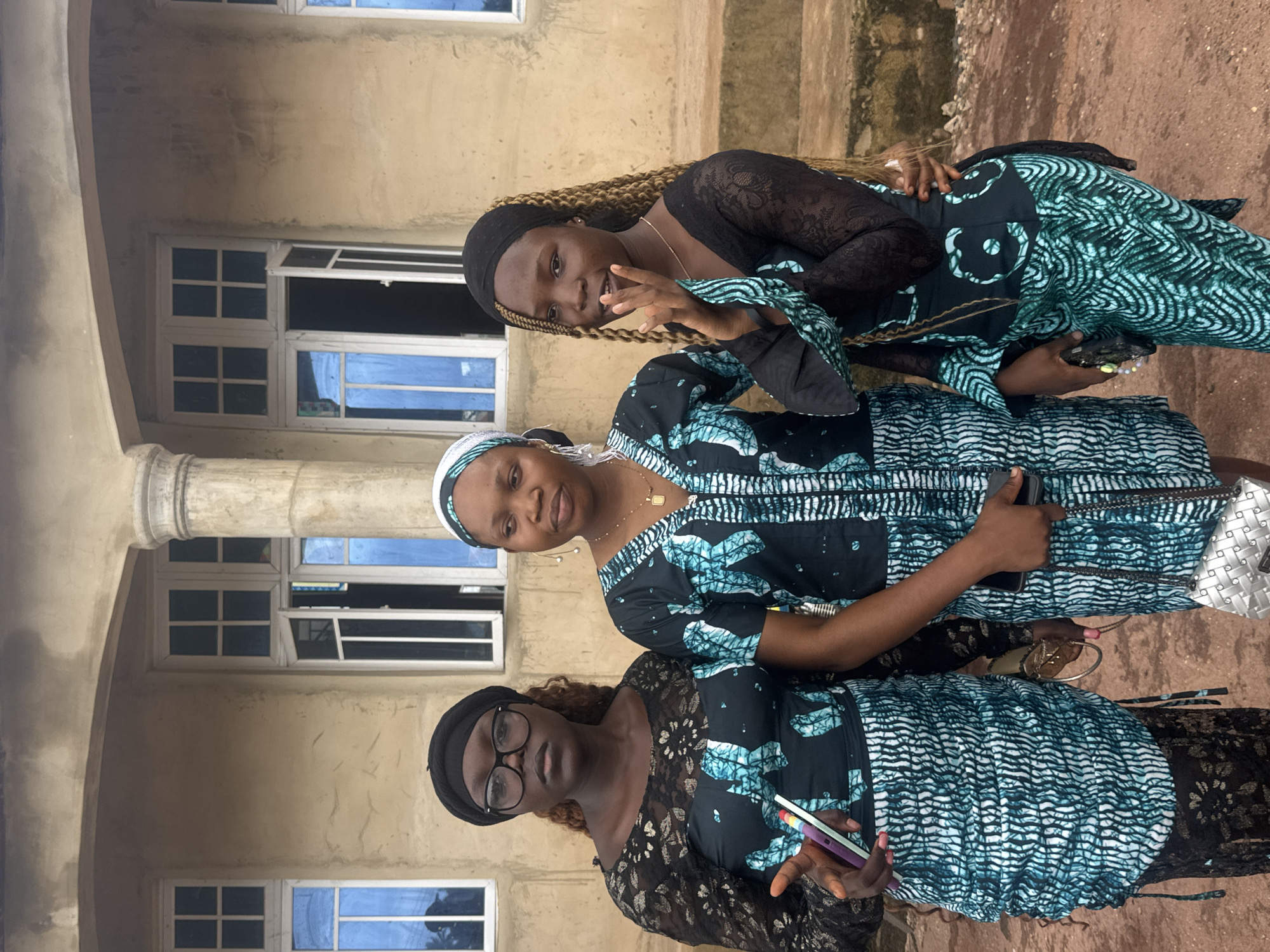 Three women stand together in front of a beige building with a column and arches in Eziudo, Nigeria. The women are dressed in similar patterned fabric with black lace accents; two have head coverings. The woman on the bottom wears glasses and holds a cell phone. The woman in the middle has her arms crossed. The woman on top is smiling and making a hand gesture. Windows with blue-tinted glass are visible behind them. The ground appears to be dirt.