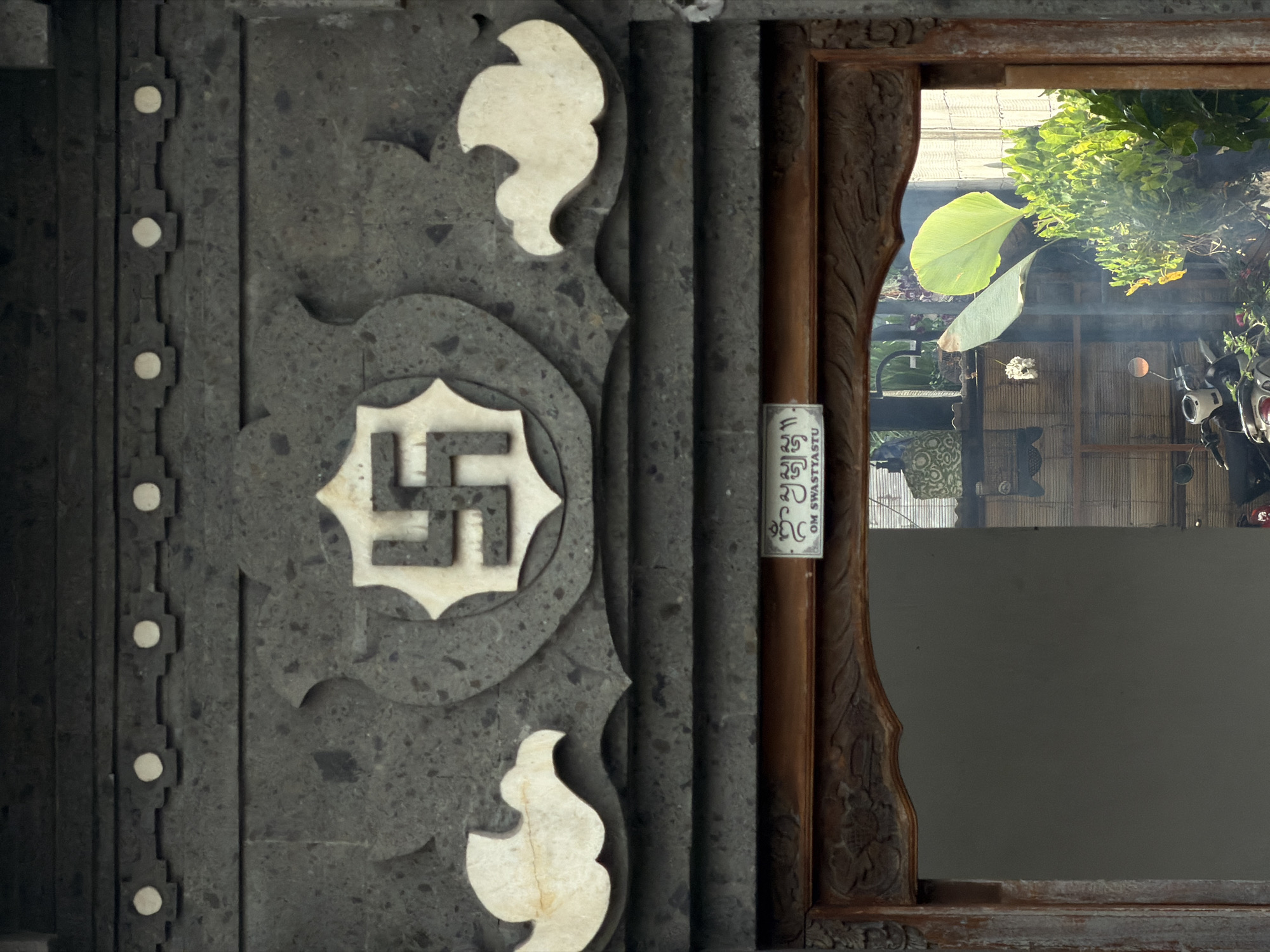 A close-up shot depicts a stone structure adorned with a swastika symbol, located in North Kuta, Indonesia. The structure is predominantly grey with white ornamental inlays. Adjacent to the stone facade is a wooden framed window, revealing a partial outdoor scene of foliage, a building, and a glimpse of a motorcycle. A small, cylindrical object with printed text is affixed near the wooden frame. The lighting appears natural, and the overall composition suggests a cultural or religious context.
