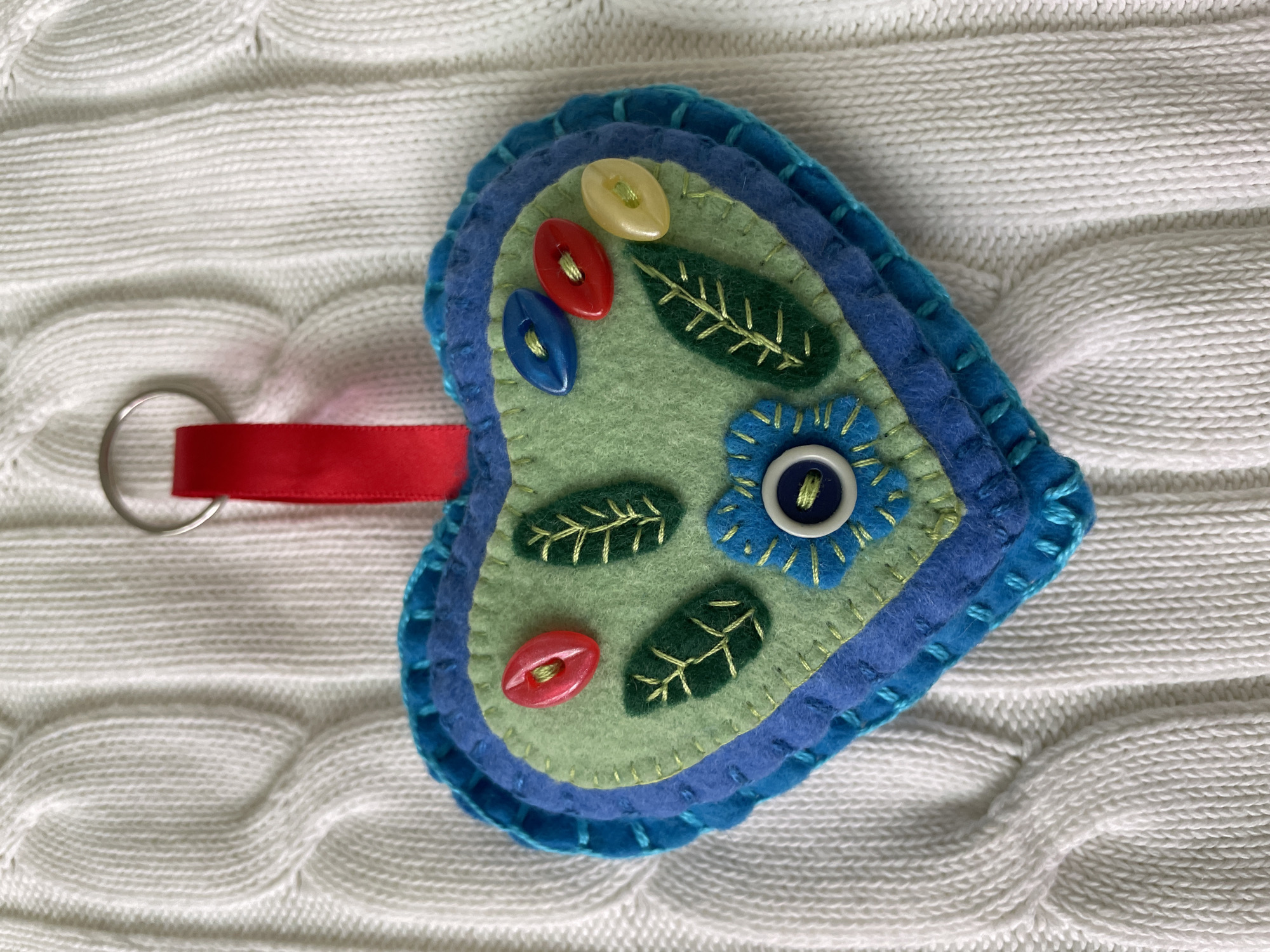 A felt heart-shaped keychain lies on a white cable-knit sweater. The keychain has layers of blue and turquoise felt, with a green felt top layer. The green layer is decorated with three oval buttons in yellow, red, and blue, three green felt leaves with yellow embroidered veins, and a blue felt flower with a blue button in the center. A silver keyring is attached to the heart by a short length of red ribbon.