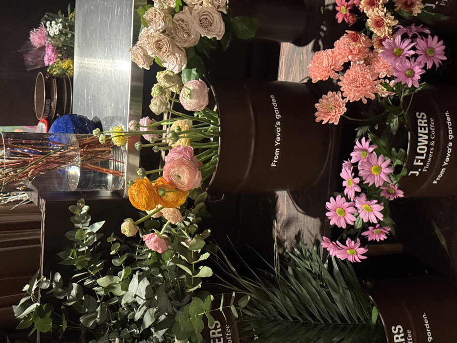A collection of flowers arranged in pots and vases. Roses, ranunculus, and daisies in shades of pink, orange, and yellow are prominent. Eucalyptus and palm leaves provide greenery. The pots have labels: "From Yeva's garden" and "J. FLOWERS Flowers & Coffee." The arrangements are placed on a shelf with a metallic surface. Some vases contain branches.
