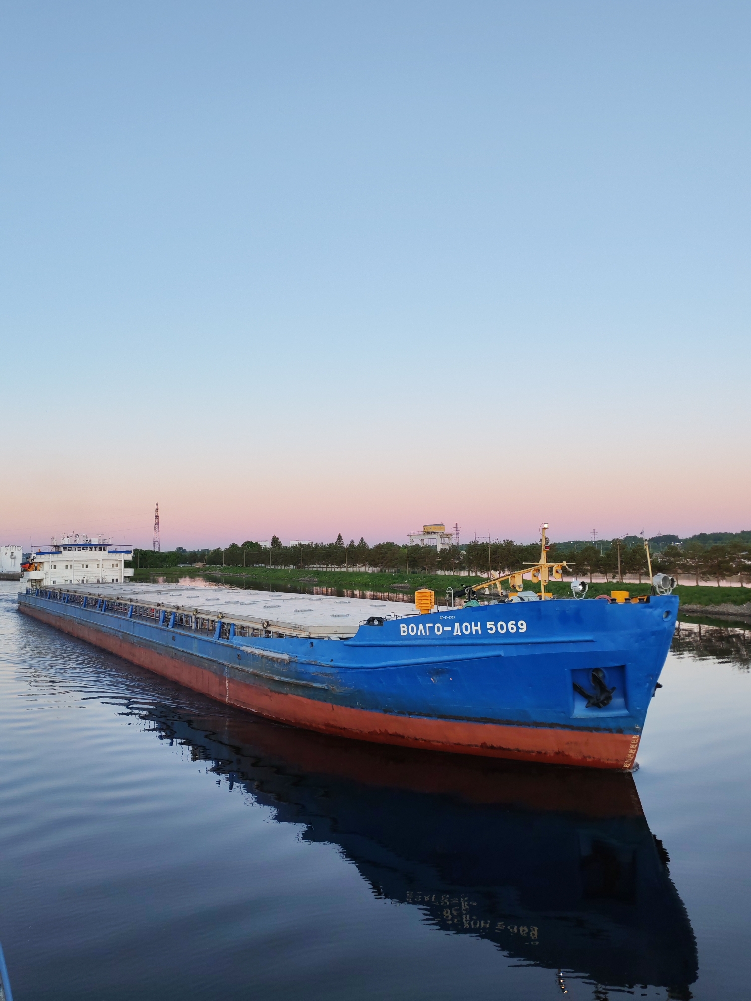 A long, blue-hulled cargo ship, identified by the Cyrillic inscription "ВОЛГО-ДОН 5069" and "ИД-21-3332" on its bow, is positioned on calm, reflective water, likely a river or canal. The ship's hull is primarily royal blue above the waterline, transitioning to a reddish-orange below, showing signs of wear and minor rust. Its superstructure, located at the stern and partially visible on the left, is white. Numerous closed cargo hatches are visible along the length of the deck. The ship's bow, facing the viewer's right, features an anchor on the port side and visible draft markings.
The sky above displays a gradient, transitioning from a light blue at the zenith to soft pinks and pale oranges near the horizon, indicative of either sunrise or sunset. The calm water perfectly reflects the ship and the sky's hues.
In the background, along the distant banks of the waterway: to the left, a white industrial building and a tall, lattice-structured tower are visible. To the right, a consistent line of green deciduous trees stretches along the shoreline, behind which faint outlines of additional distant structures are discernible. No human figures are present, and the ship appears to be stationary or moving slowly given the minimal wake.