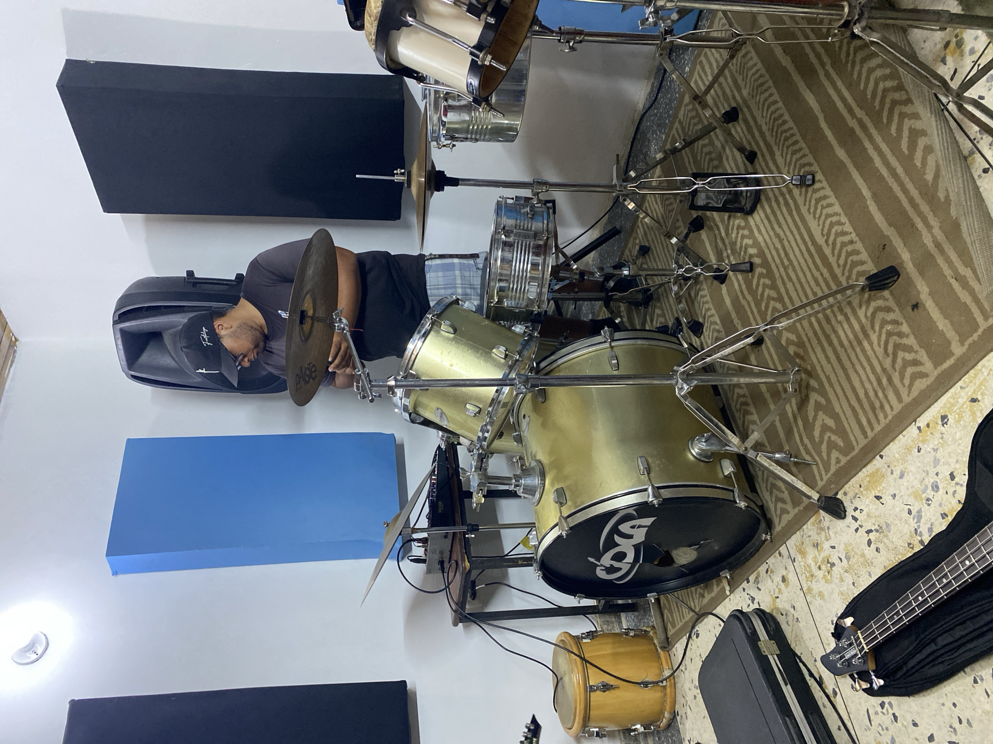 The image depicts an indoor music studio located in Tinaquillo, Venezuela. A male individual, wearing a dark t-shirt, plaid shorts, eyeglasses, and a dark baseball cap, stands centrally, adjusting a gold-colored cymbal on a drum stand. He is positioned behind a gold-shelled drum kit, which includes a large bass drum with a black front head displaying a white "DDCA" logo and a chrome snare drum.
The studio features white walls with multiple sound-absorbing panels: two black rectangular panels are on the left and right, and one prominent blue rectangular panel is central on the left wall. A bright white circular light fixture is mounted high on the upper left wall. The floor is covered with light-colored, speckled tiles, over which a rectangular brown and beige patterned rug is laid beneath the drum kit.
Additional musical equipment includes a light wood-colored conga drum to the far left. Behind the conga, a small dark wooden table holds electronic audio equipment, including a small mixer or multi-track recorder with multiple cables connected. A large, dark grey speaker cabinet is positioned behind the individual. In the lower left foreground, an open black hardshell case contains a black bass guitar, partially visible within a soft case. Various chrome drum stands with black rubber feet are distributed across the rug.